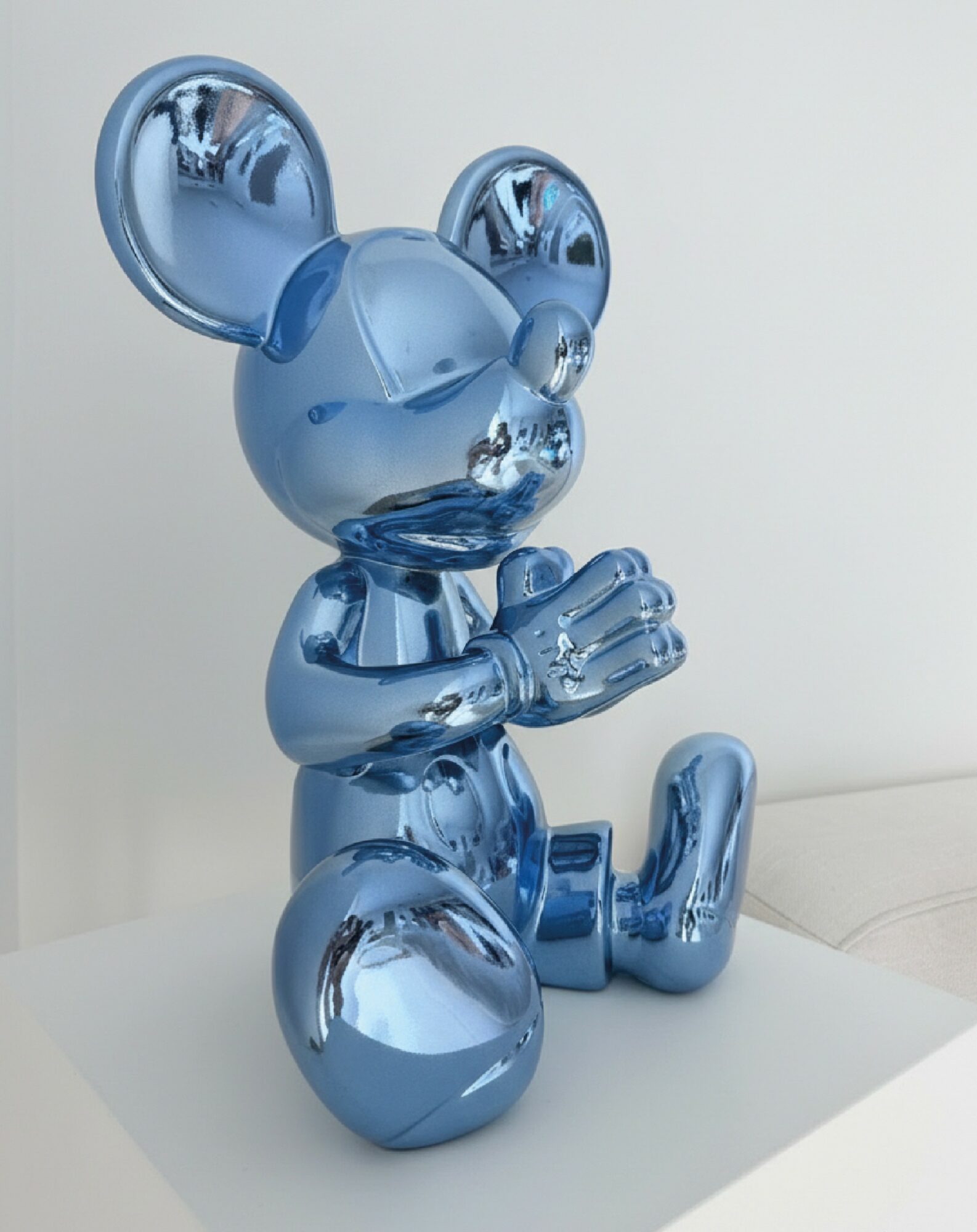 Blue metallic mouse sculpture sitting with hands together, on a white surface, against a plain background.