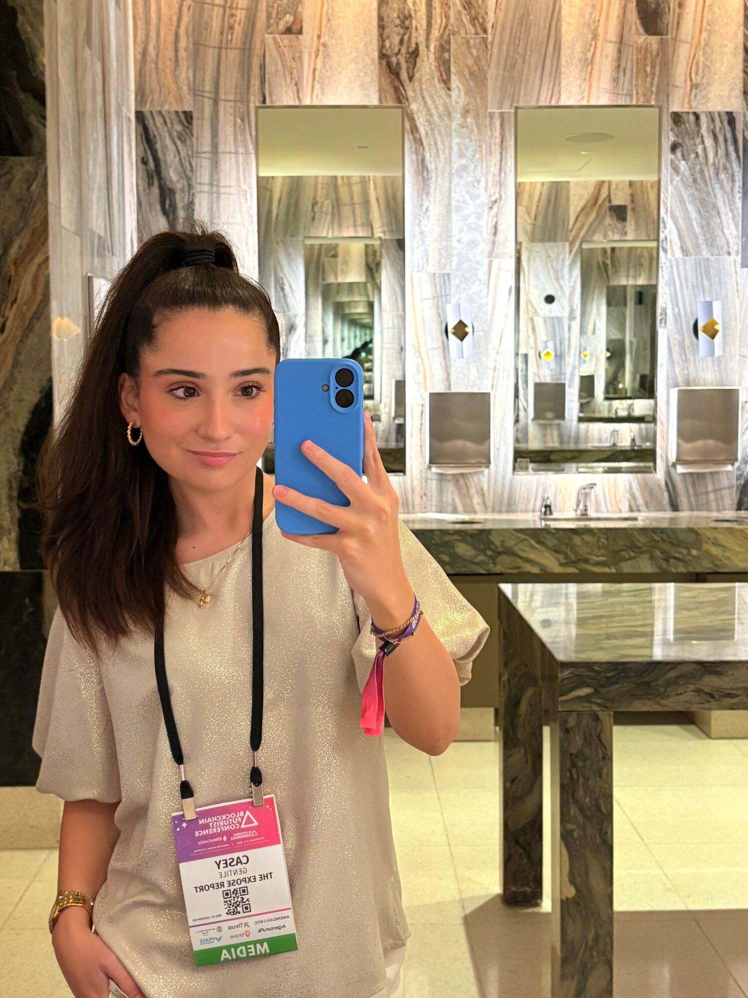 Young woman with dark hair in a ponytail taking a mirror selfie in a room with marble walls and mirrors, wearing a beige top and a pink badge.