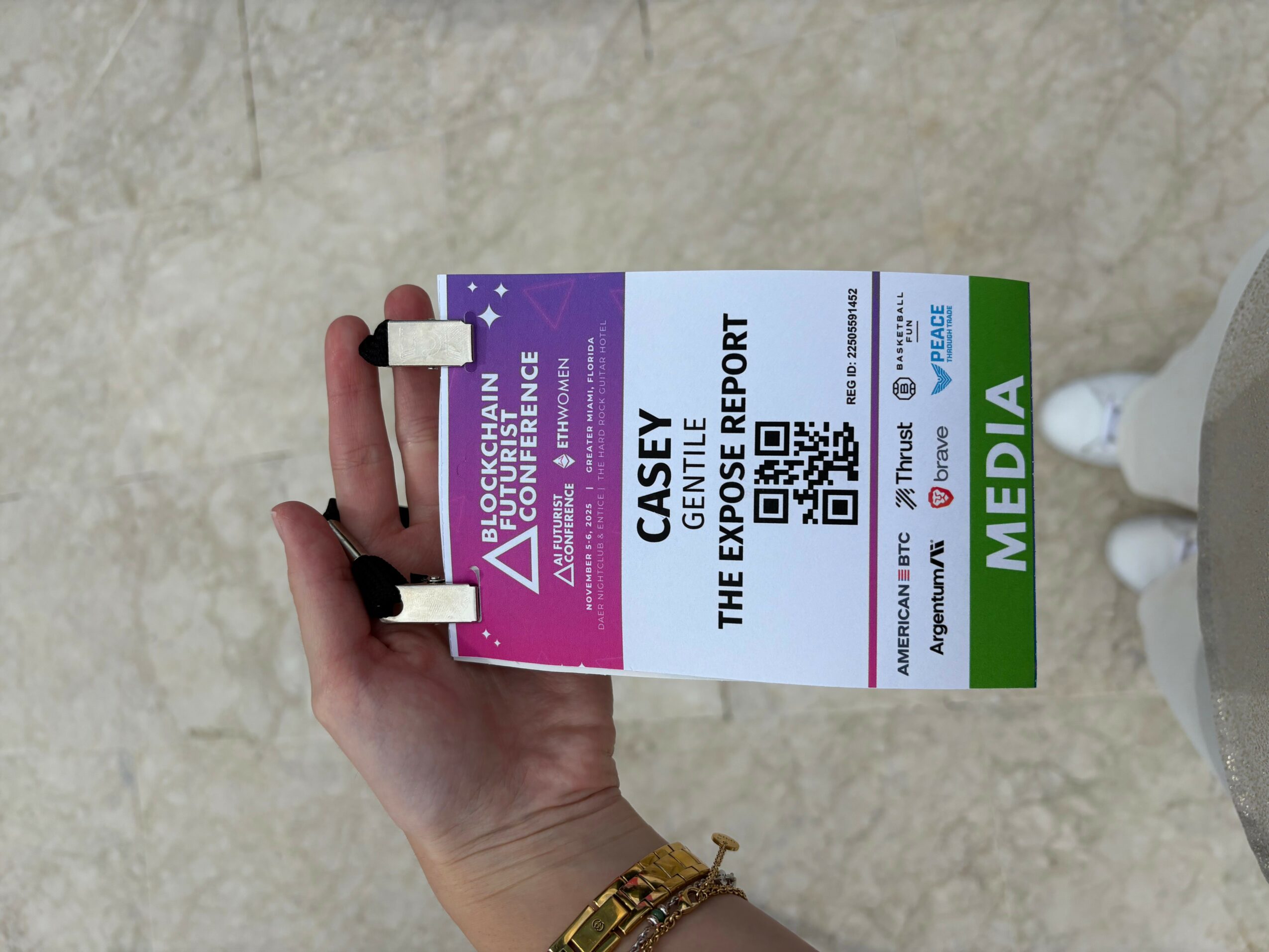 Hand holding a media pass with purple, white, and green sections, displaying a QR code and text, against a beige background.