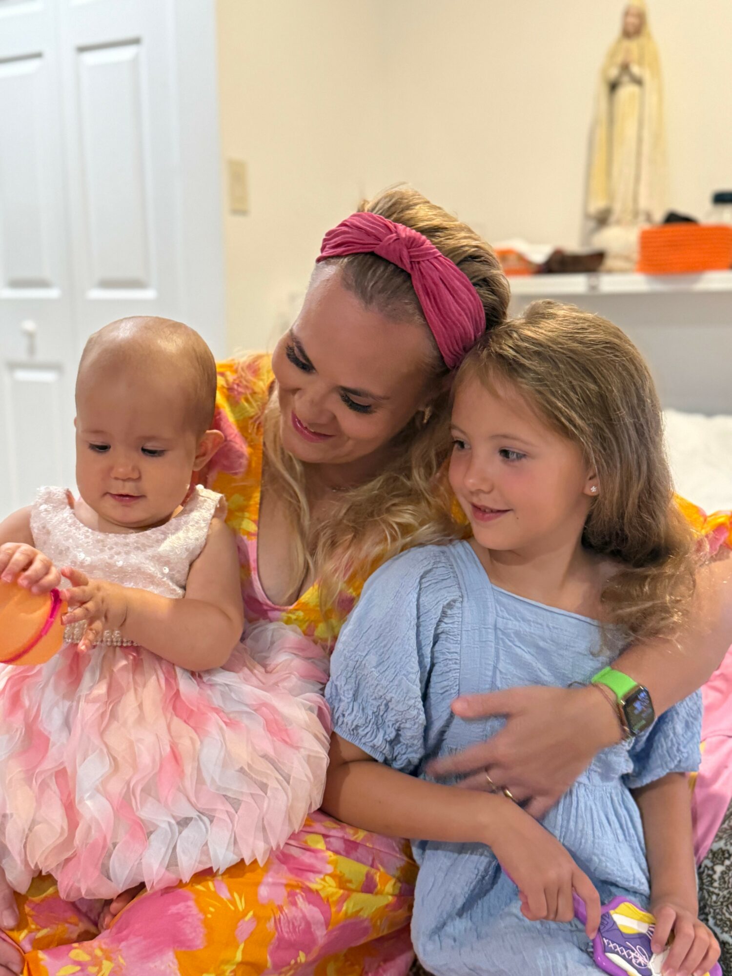 Woman with two children, one in a pink dress, in a room with a statue and a table, smiling and hugging.