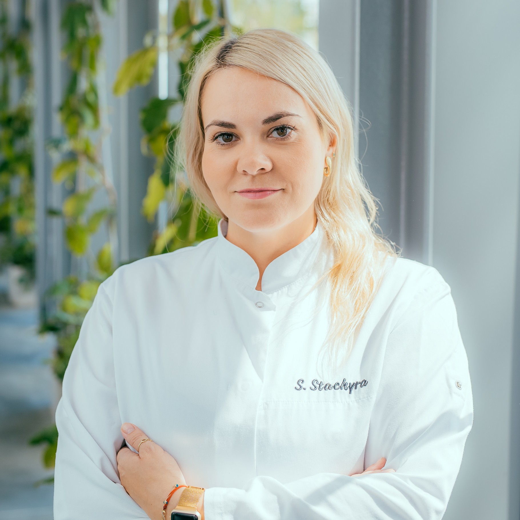 Woman in white chef's coat with name embroidered, standing indoors near window with plants, arms crossed, looking at camera.
