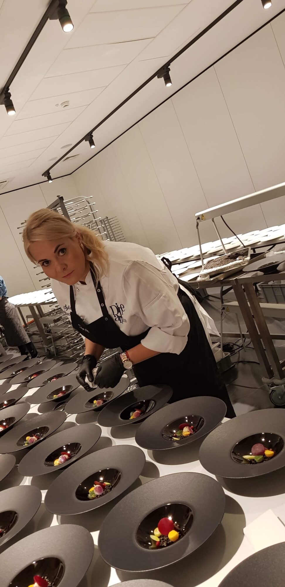 Person with blonde hair wearing a white shirt and black apron working with circular objects on a table.
