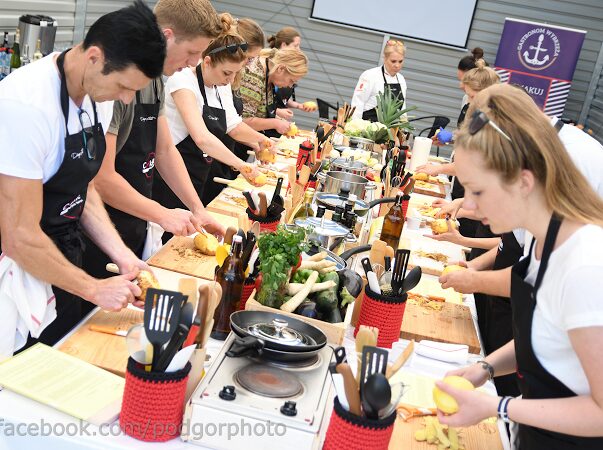 People participating in a cooking class outdoors, preparing food at a long table with cooking utensils and ingredients.