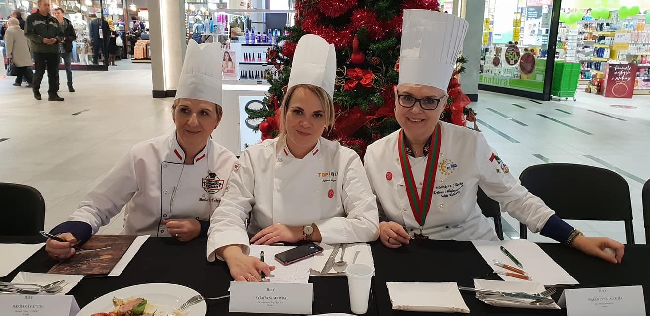 Four children dressed as chefs sit at a table with a Christmas tree behind them, in a shopping mall. They wear chef hats and uniforms.