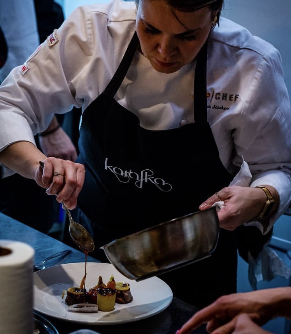 Chef drizzling sauce onto plated desserts in a professional kitchen, wearing a white uniform and black apron.