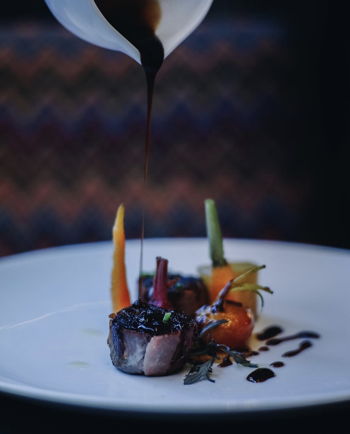 Chef drizzles dark sauce over a plated gourmet dish with colorful vegetables and a piece of meat on a white plate.