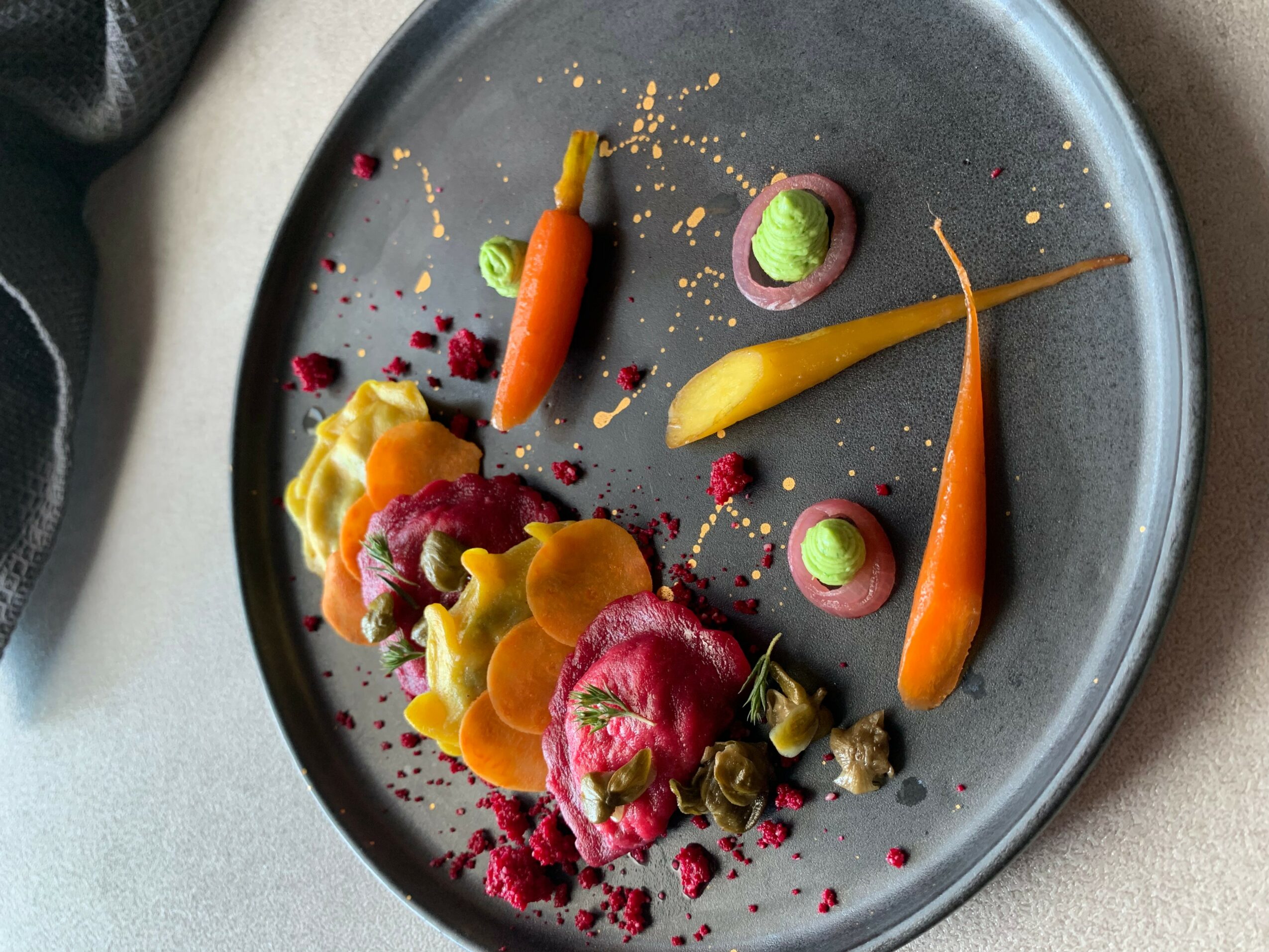 Assorted colorful vegetables and garnishes arranged on a dark plate, with some scattered crumbs and sauce, viewed from above.