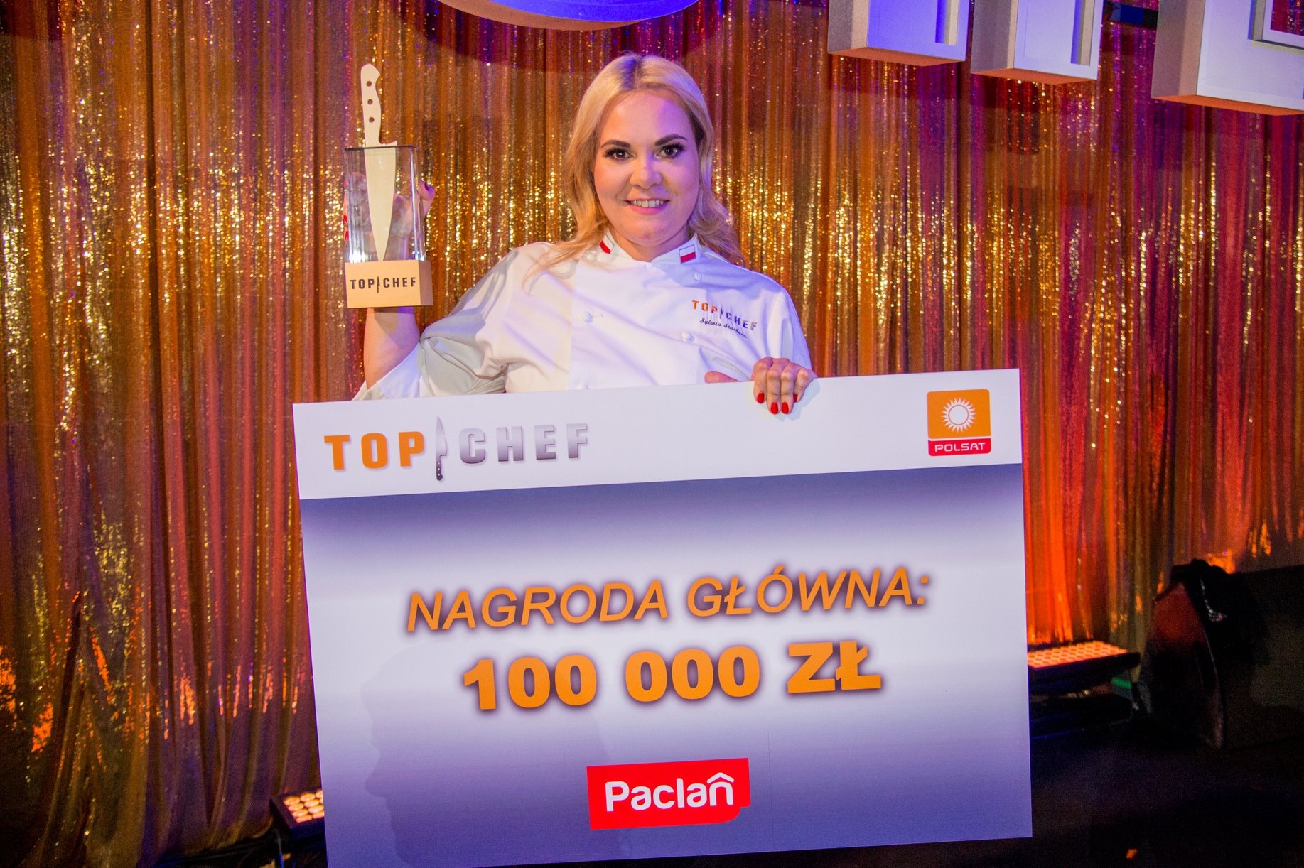 Woman holding a large check for 100,000 zł on stage with a gold curtain background.