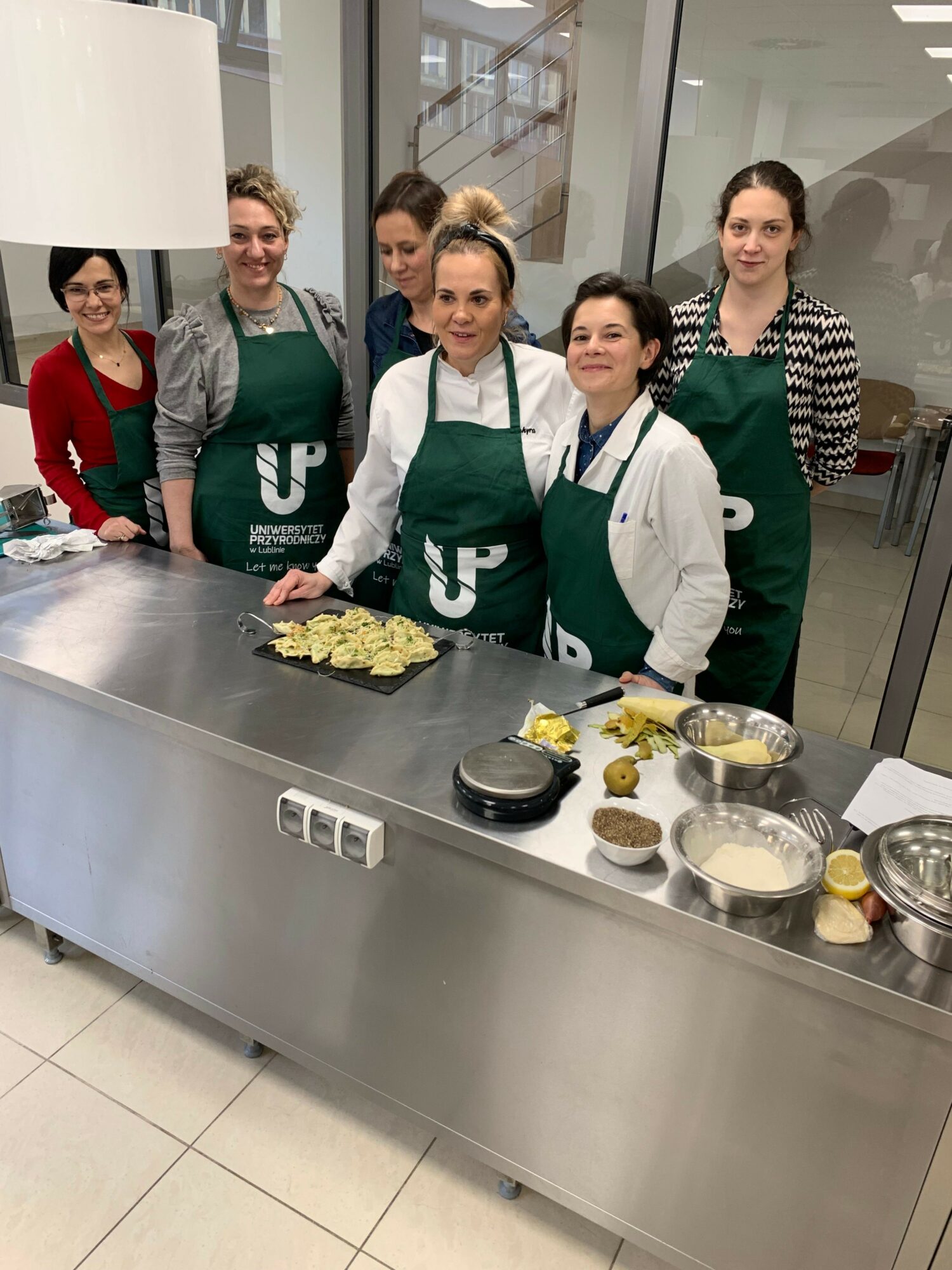 Four women in aprons and chef jackets stand behind a stainless steel kitchen counter with food ingredients and a cake, smiling.