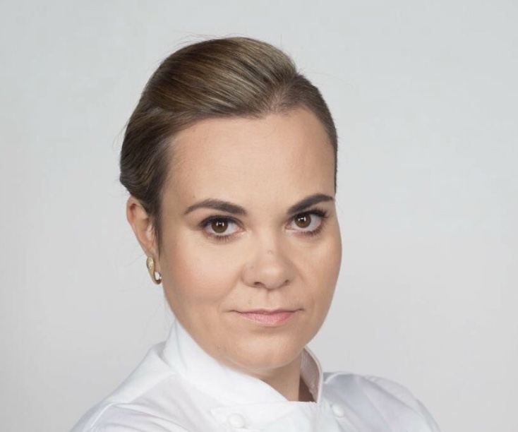 Portrait of a woman with brown hair styled back, wearing a white top, looking directly at the camera.