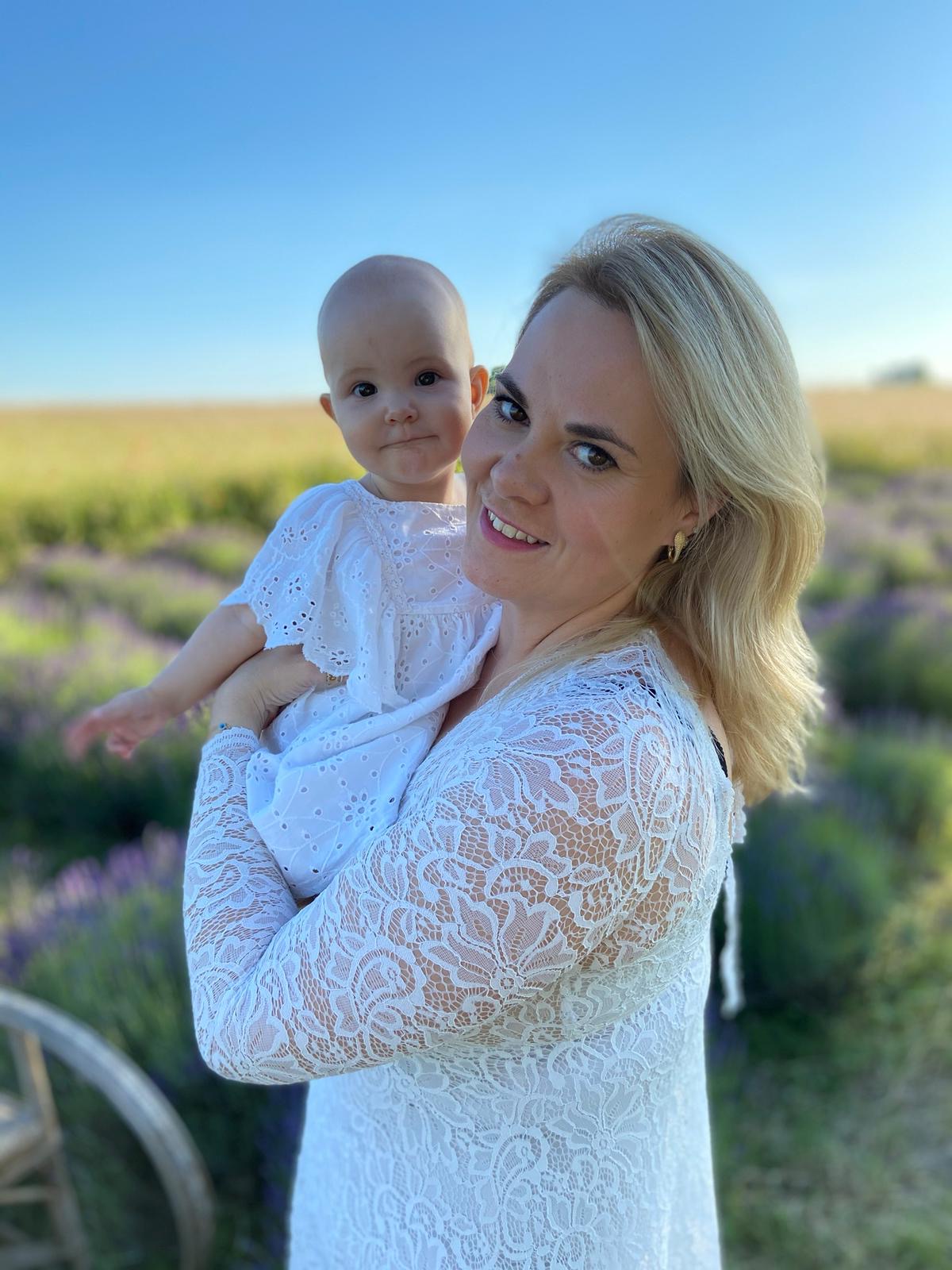 Woman holding a baby outdoors in a field with a clear blue sky, both smiling, woman wearing a white lace top.