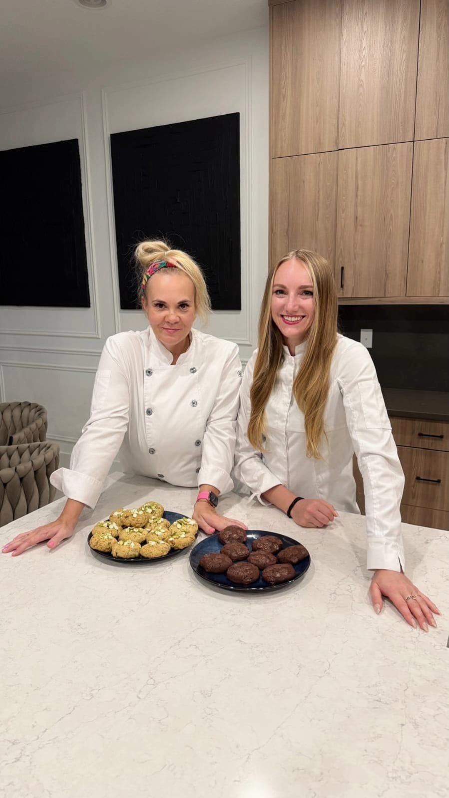 Two women in white chef jackets stand behind a kitchen counter with plates of cookies, smiling.