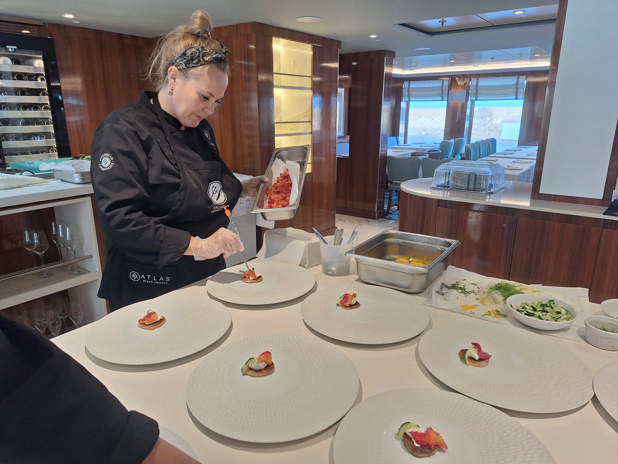 Chef preparing food in a kitchen with a dining area in the background, six empty plates with small food garnishes, and various ingredients on the counter.