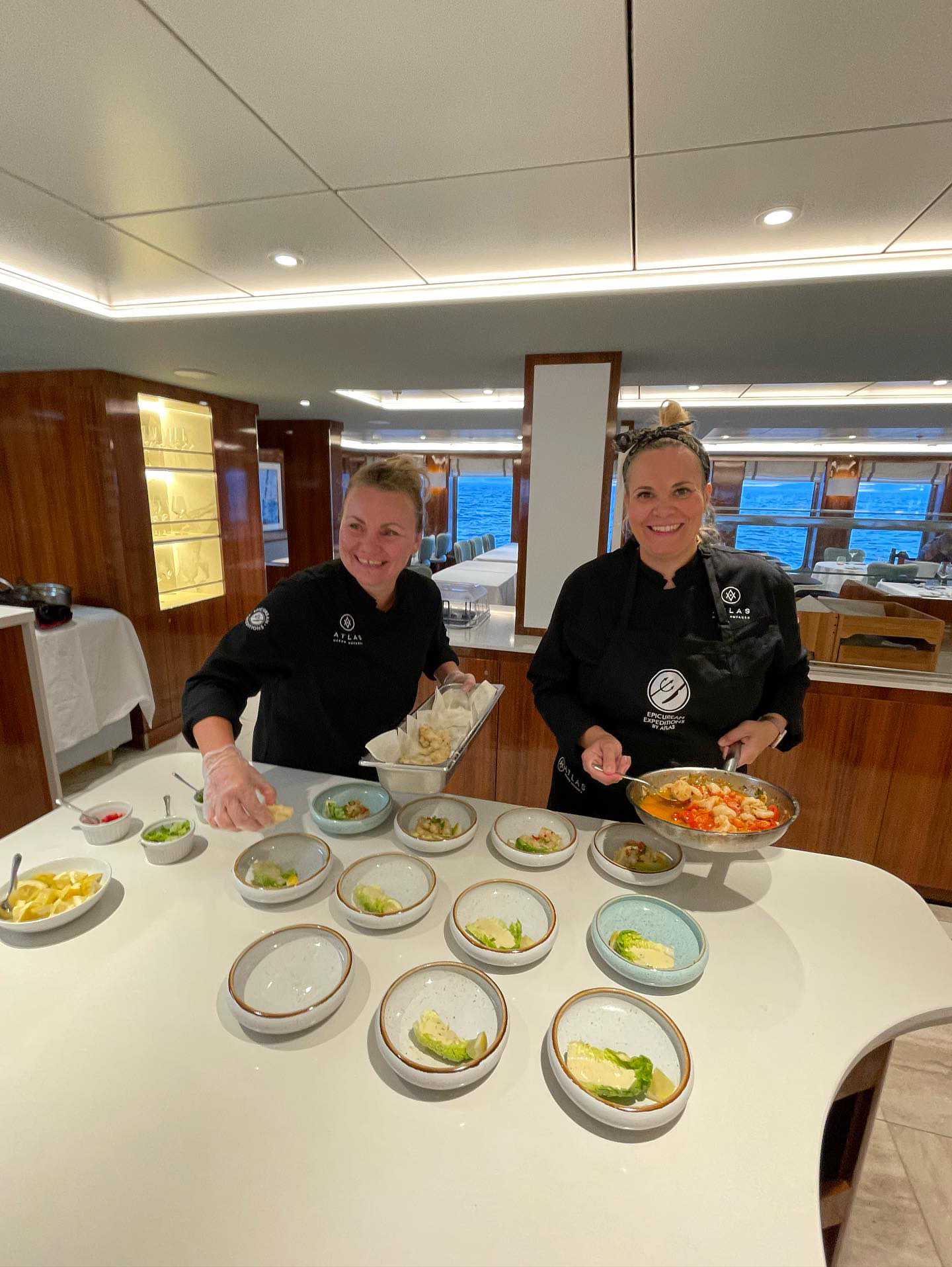 Two women in black uniforms stand behind a table with small bowls of food, smiling in a room with large windows.