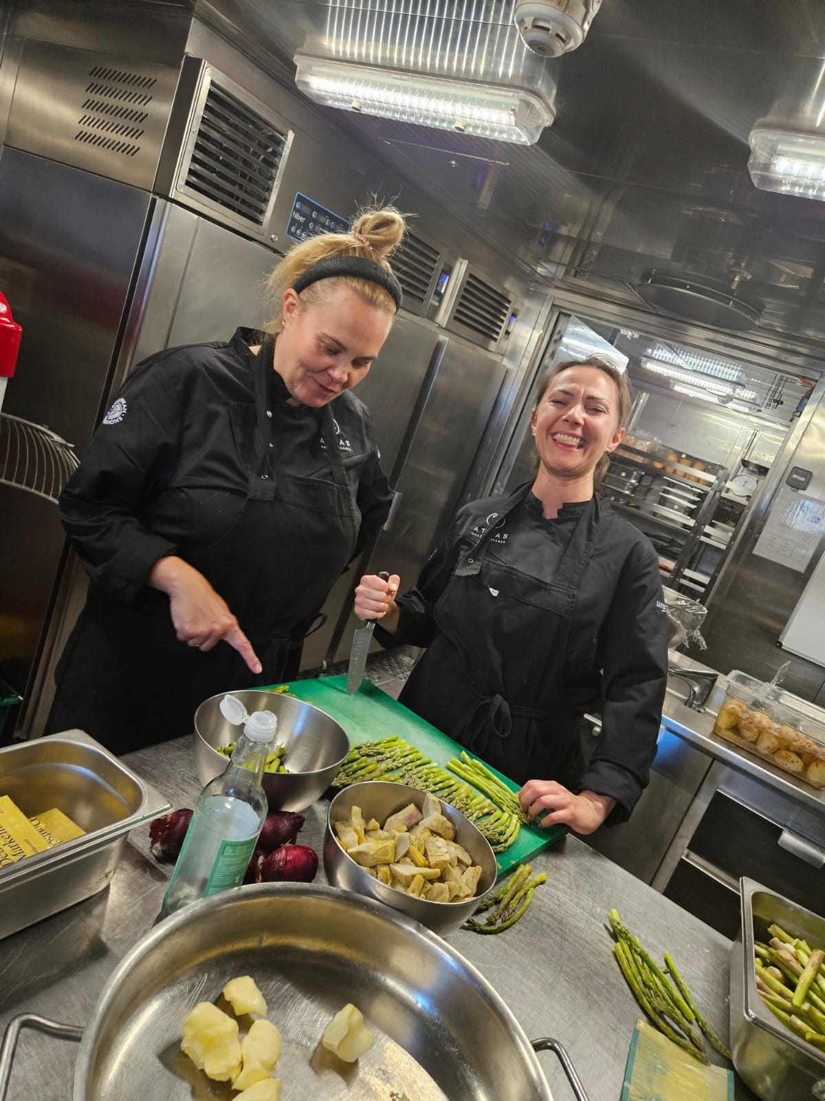 Two women in black chef jackets smiling and talking in a commercial kitchen with stainless steel appliances and ingredients on the counter.