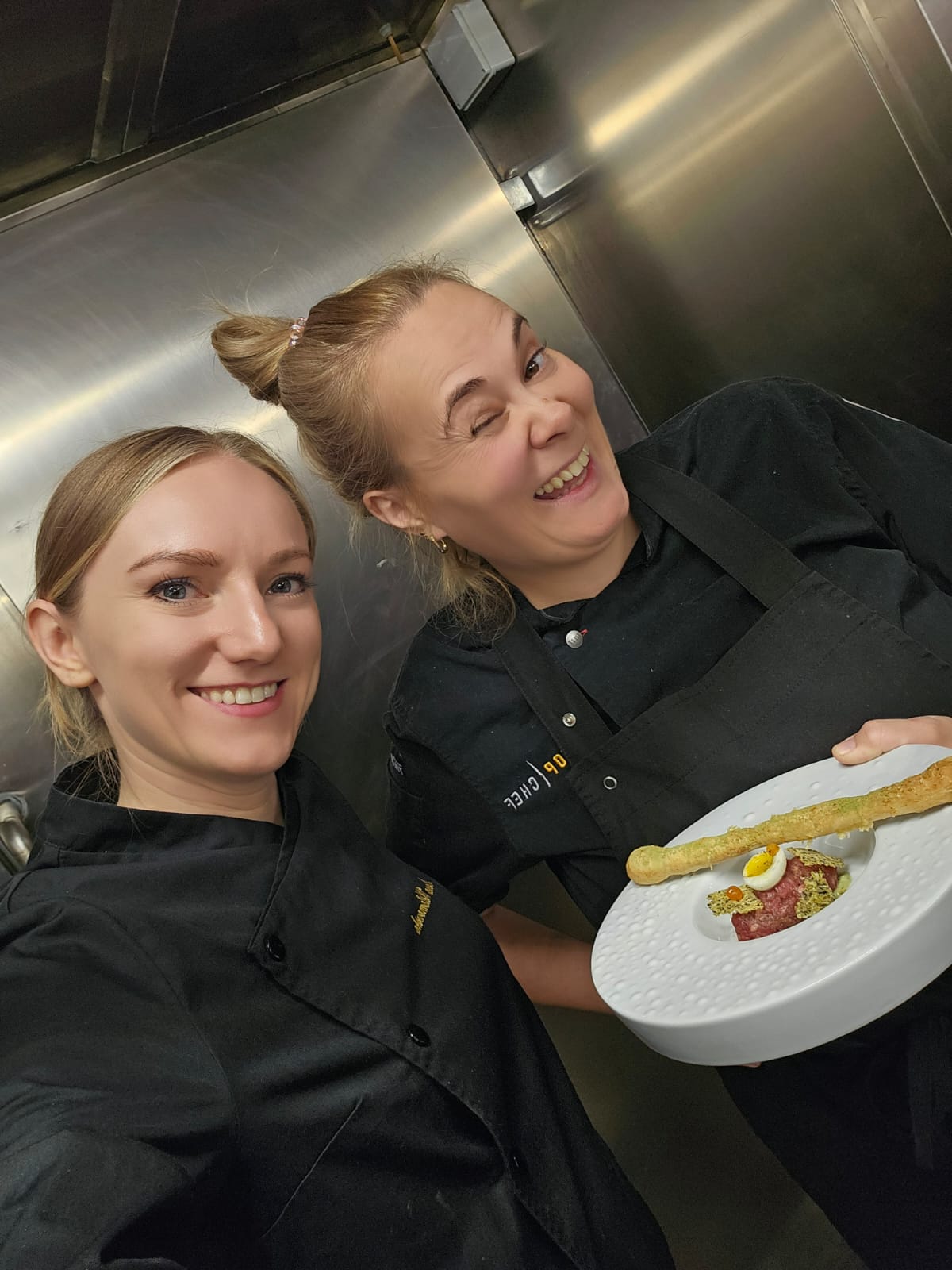Two women smiling, one holding a plate with food, in a metallic kitchen environment.