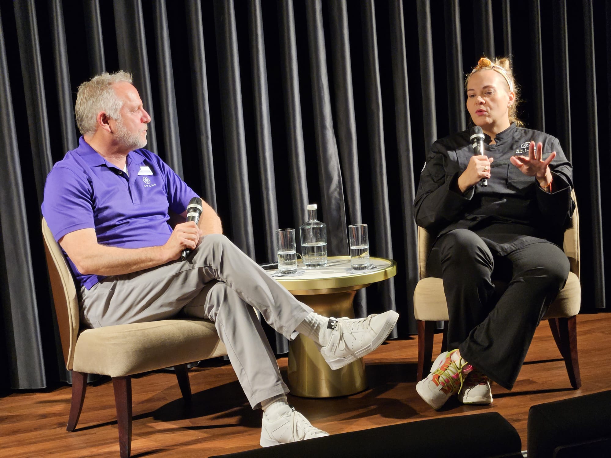 Two people seated on stage, one holding a microphone, engaged in conversation with a black curtain backdrop.