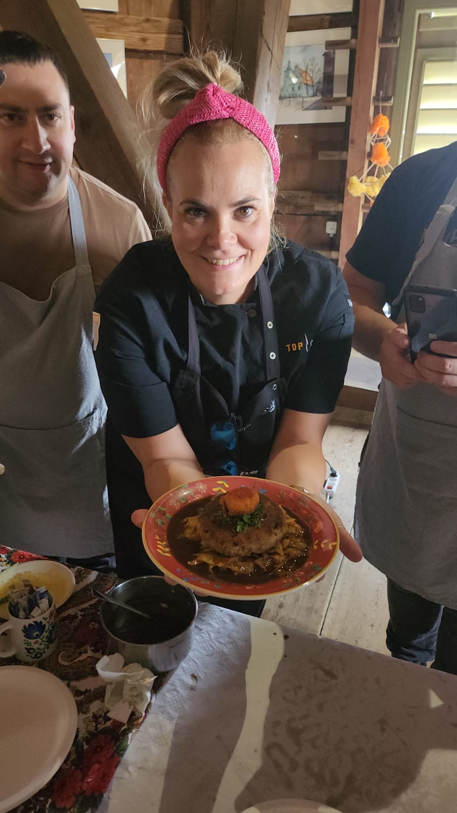 Woman with pink headband holding a plate of food, surrounded by people in a kitchen setting.