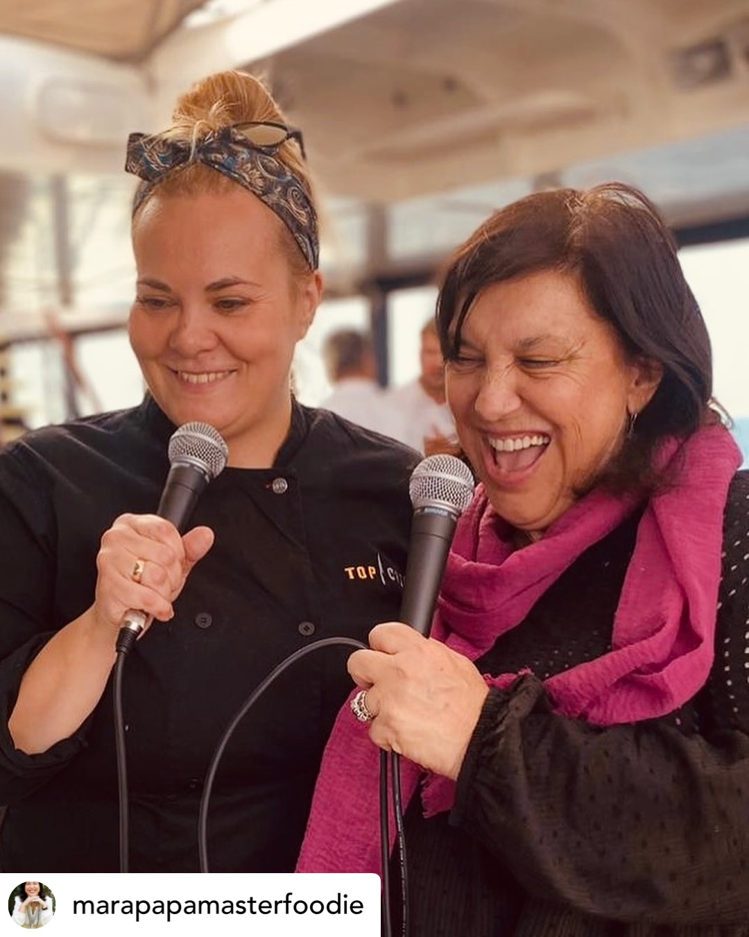 Two women smiling and holding microphones indoors, one wearing a black shirt and the other a pink scarf.