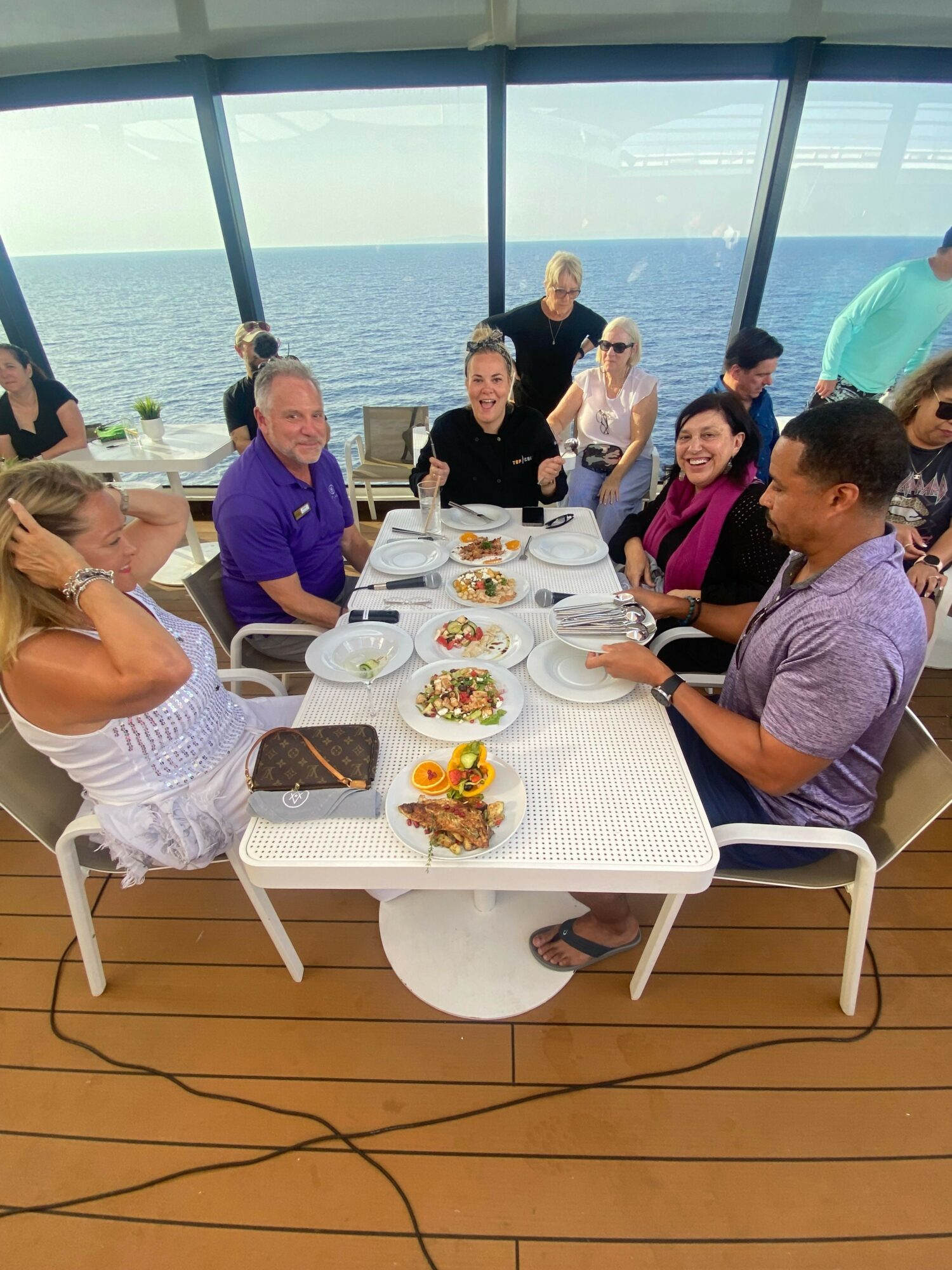 People gathered around a table with food, on a balcony overlooking water, with a view of the city skyline.
