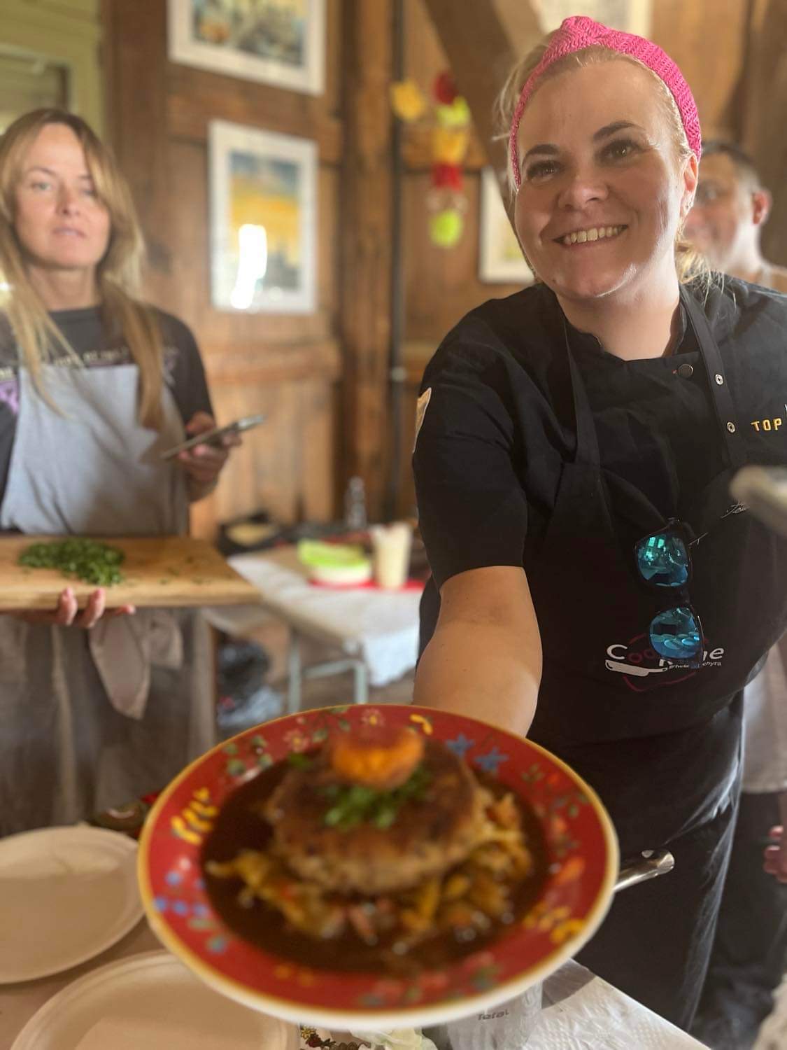 Smiling woman with pink headband presenting a dish on a red plate in a restaurant setting.