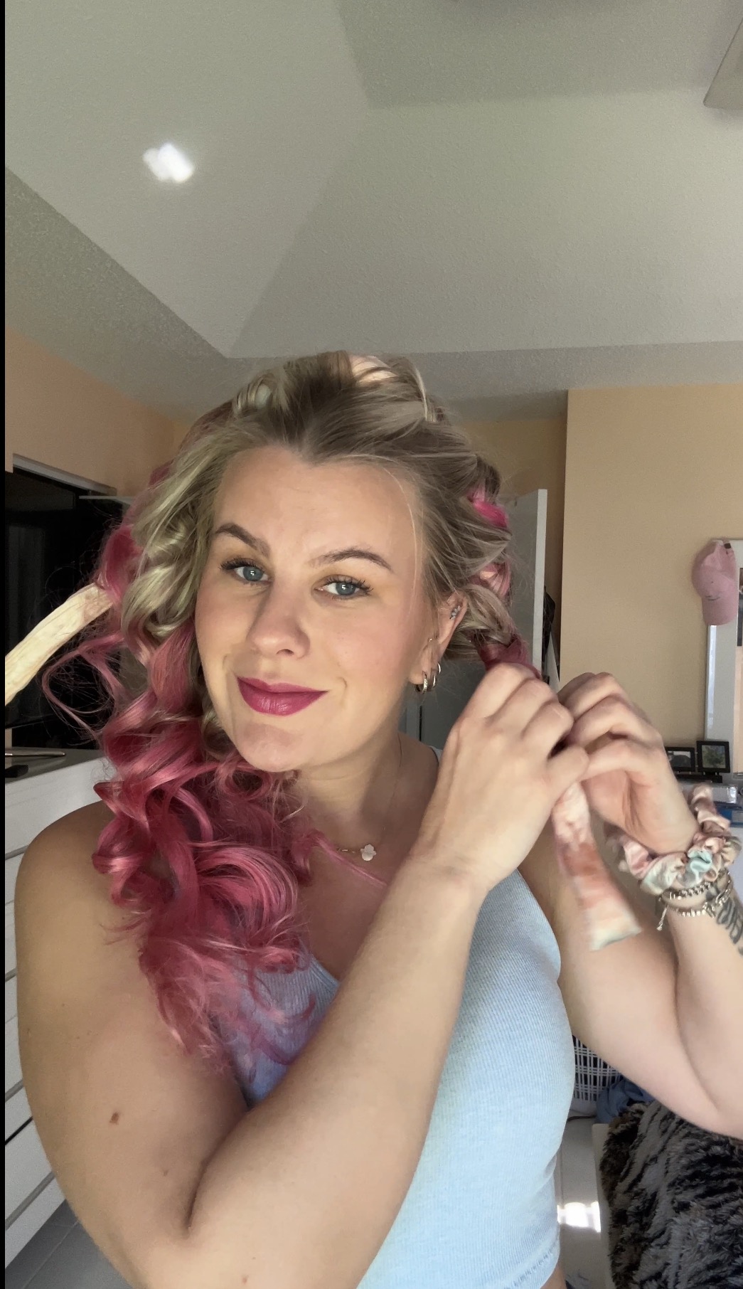 Woman with pink and blonde curly hair, wearing a light blue top, smiling, indoors.