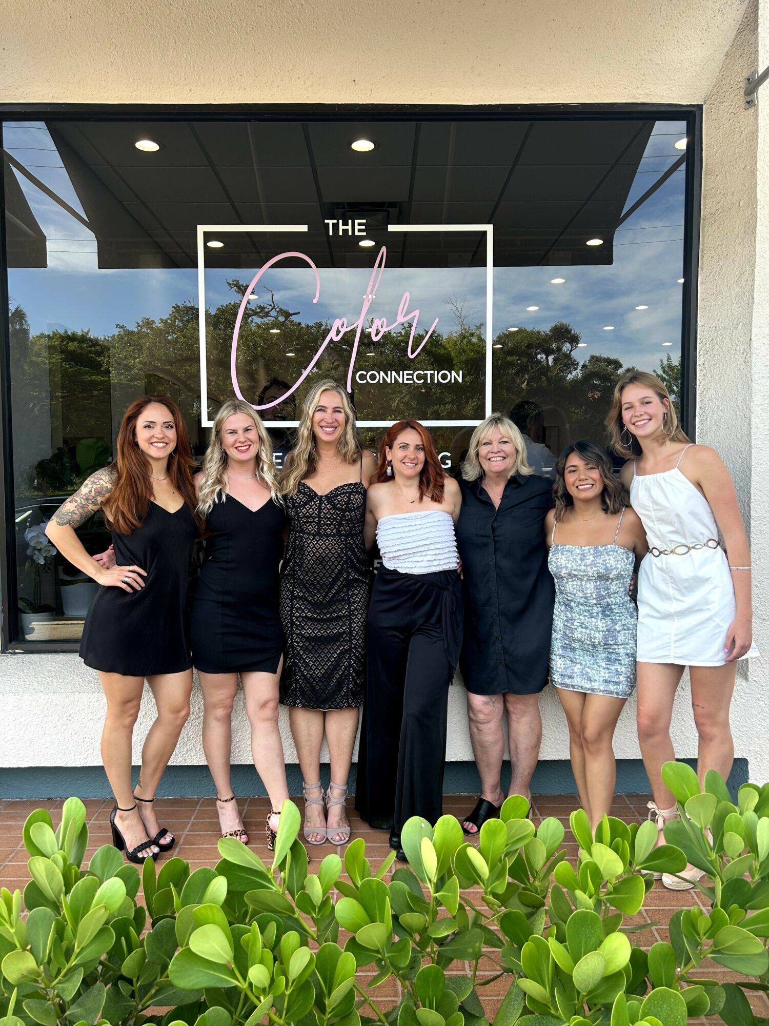 Seven women standing in front of a glass storefront with a sign reading 'The Color Connection,' smiling, with greenery in the foreground.