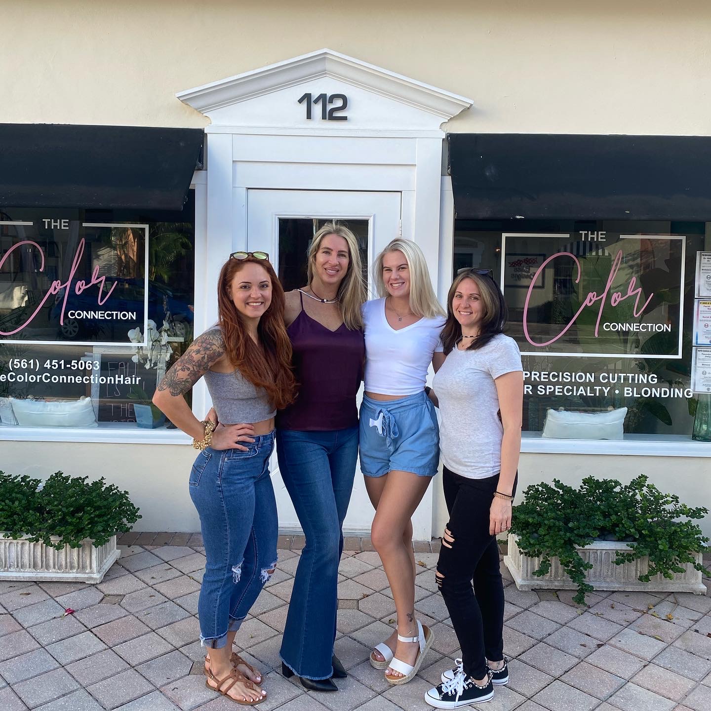 Four women stand in front of a storefront with the number 112 above the door, smiling and posing together.