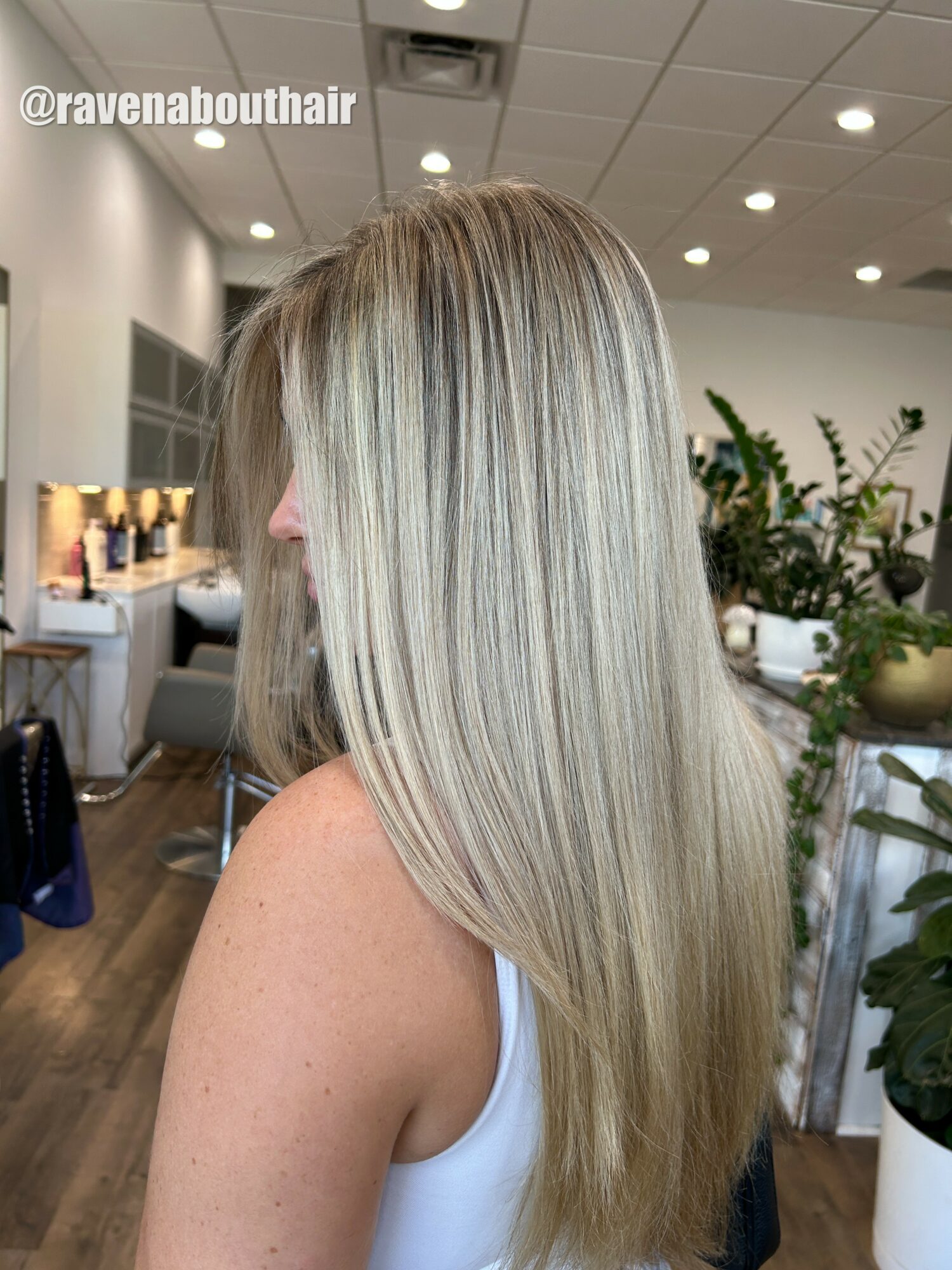 Side view of a woman with long, straight, blonde hair in a salon, with plants and salon equipment in background.
