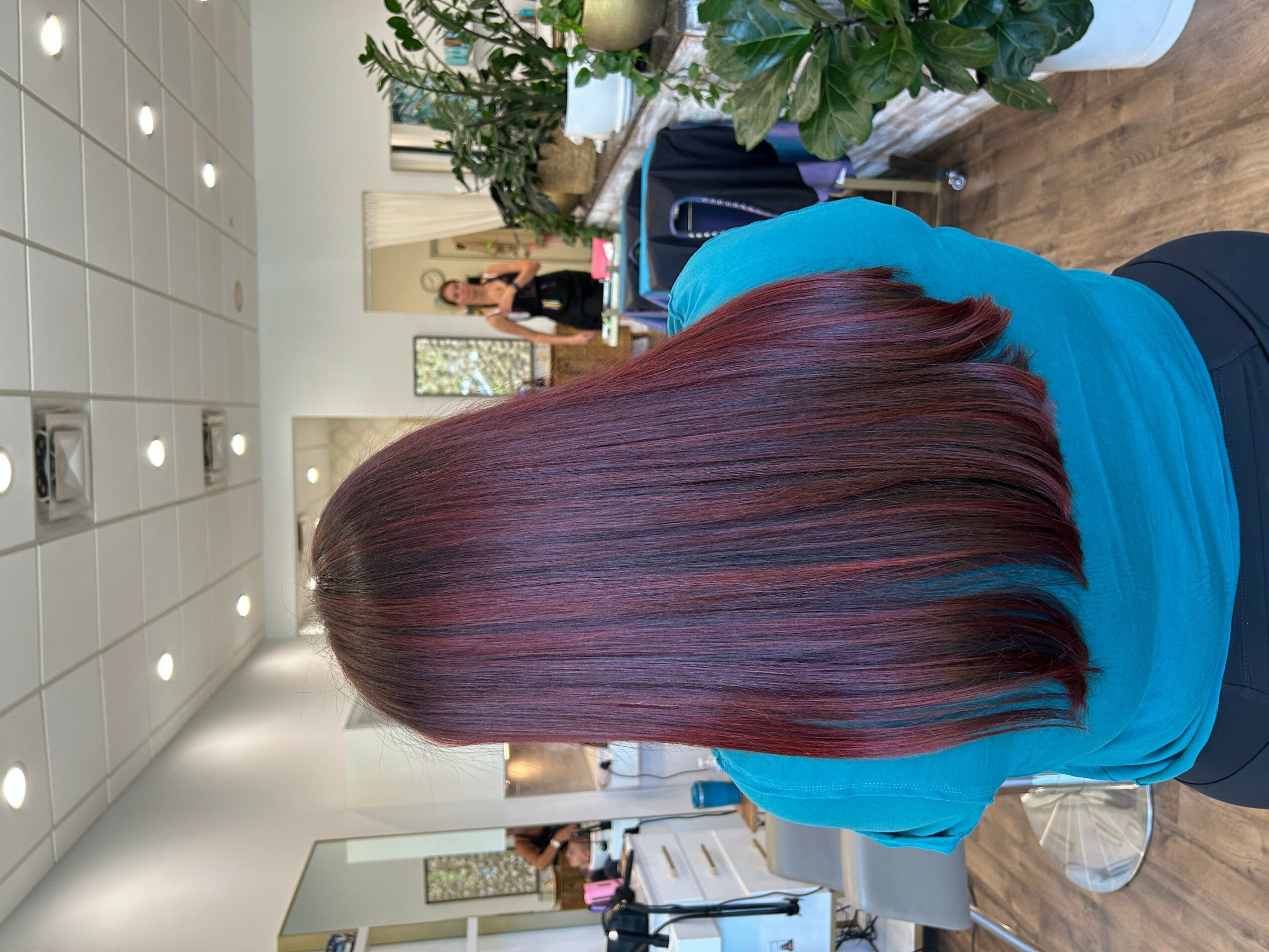 Back view of a person with long, straight, burgundy-colored hair in a salon, wearing a blue top.
