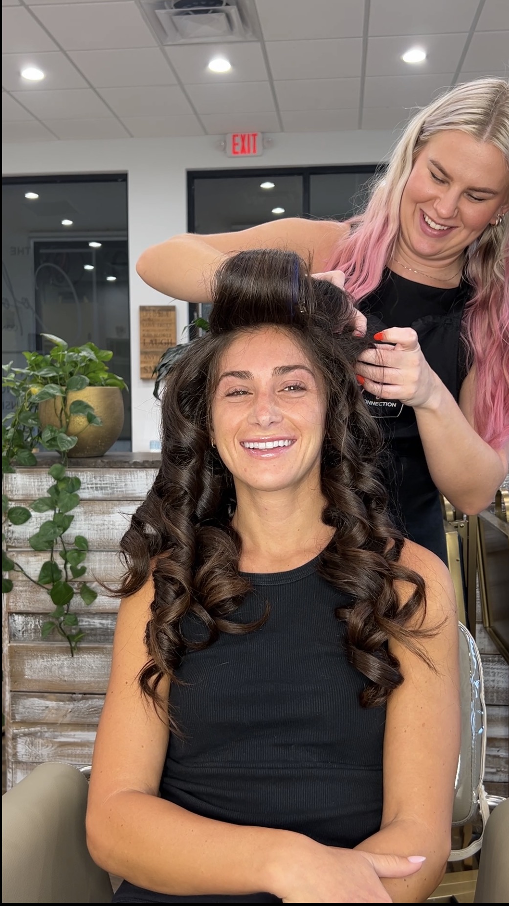 Woman with long curly hair smiling, sitting in a salon chair, hairstylist with pink hair styling her hair, indoor setting
