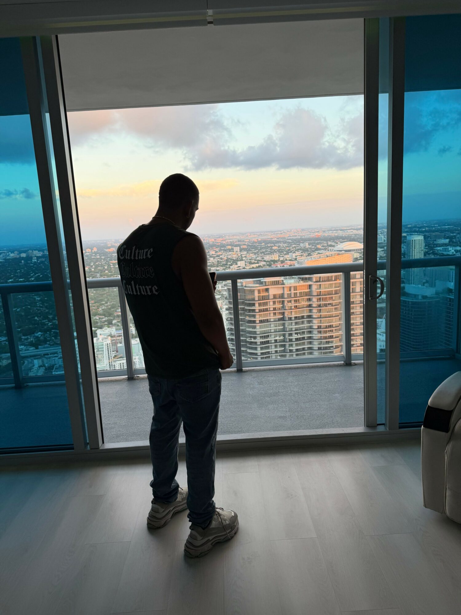 Person standing indoors near large window with city view, facing sideways, silhouette visible, sunset sky outside.
