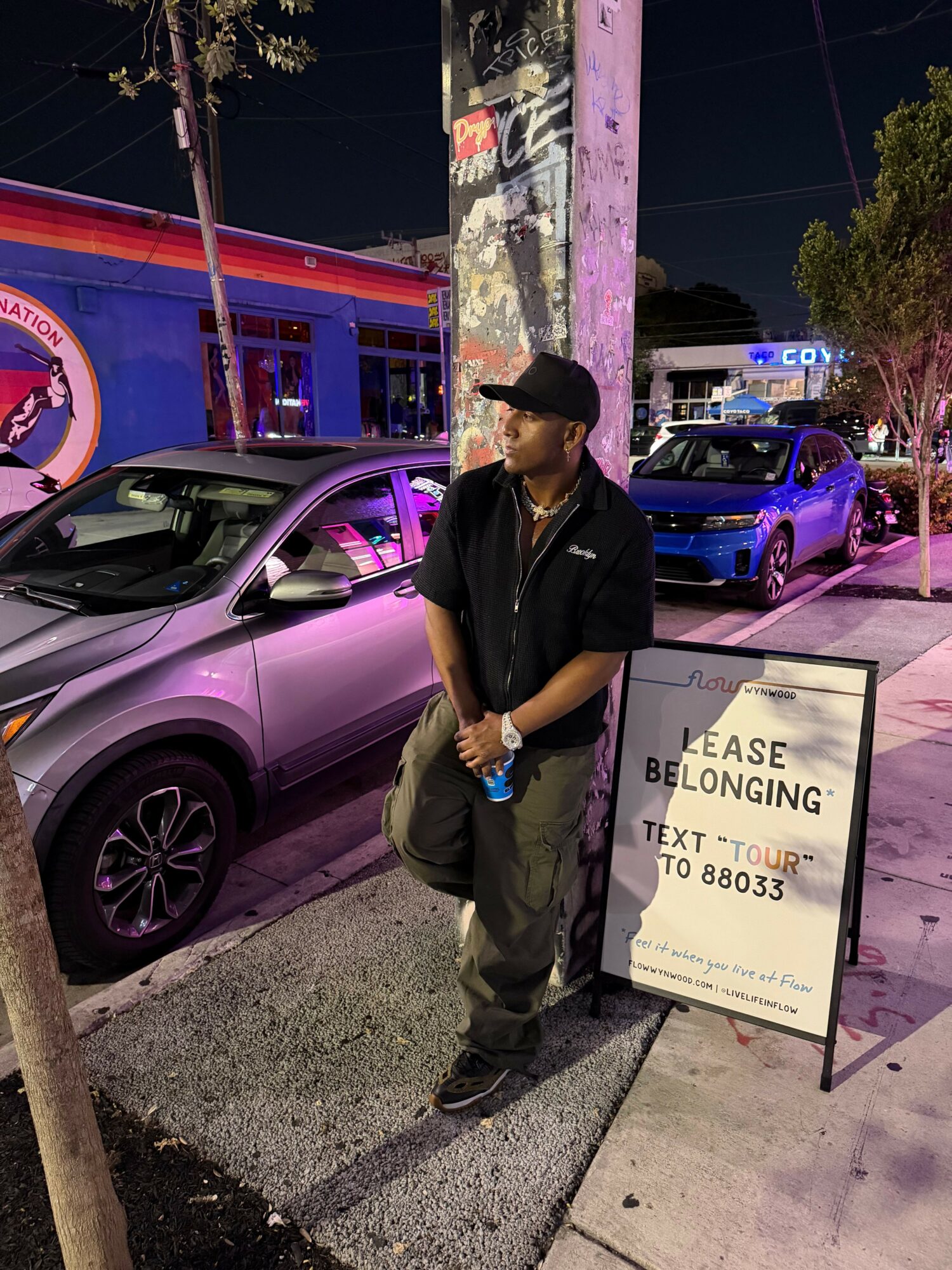 Person standing next to a purple car on a city street at night, with a sign and police car in background.