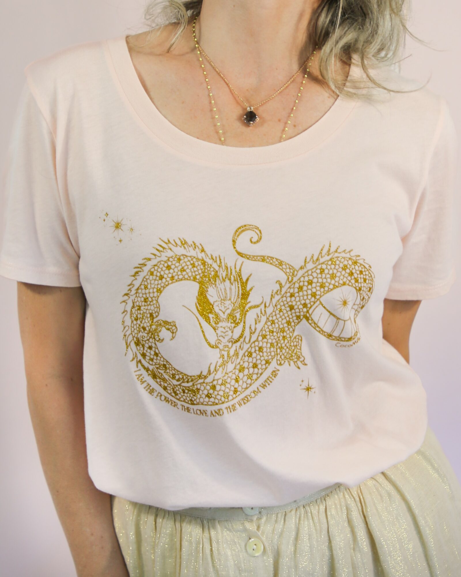 Person wearing a white T-shirt with a gold dragon design, gold necklace, and light-colored pants.