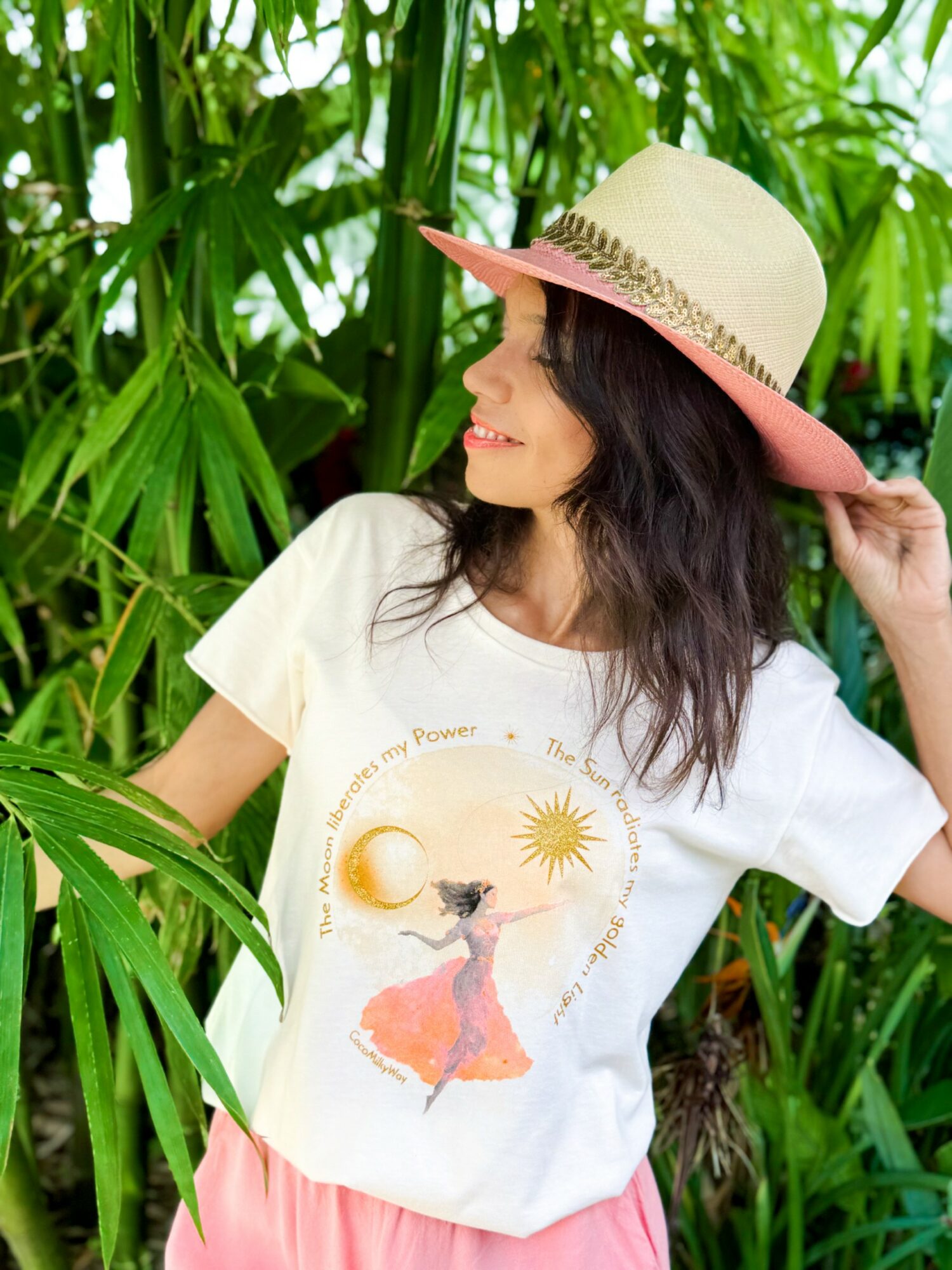 Woman wearing a wide-brimmed hat and white t-shirt with a sun and figure design, standing among green plants, smiling and looking to the side.