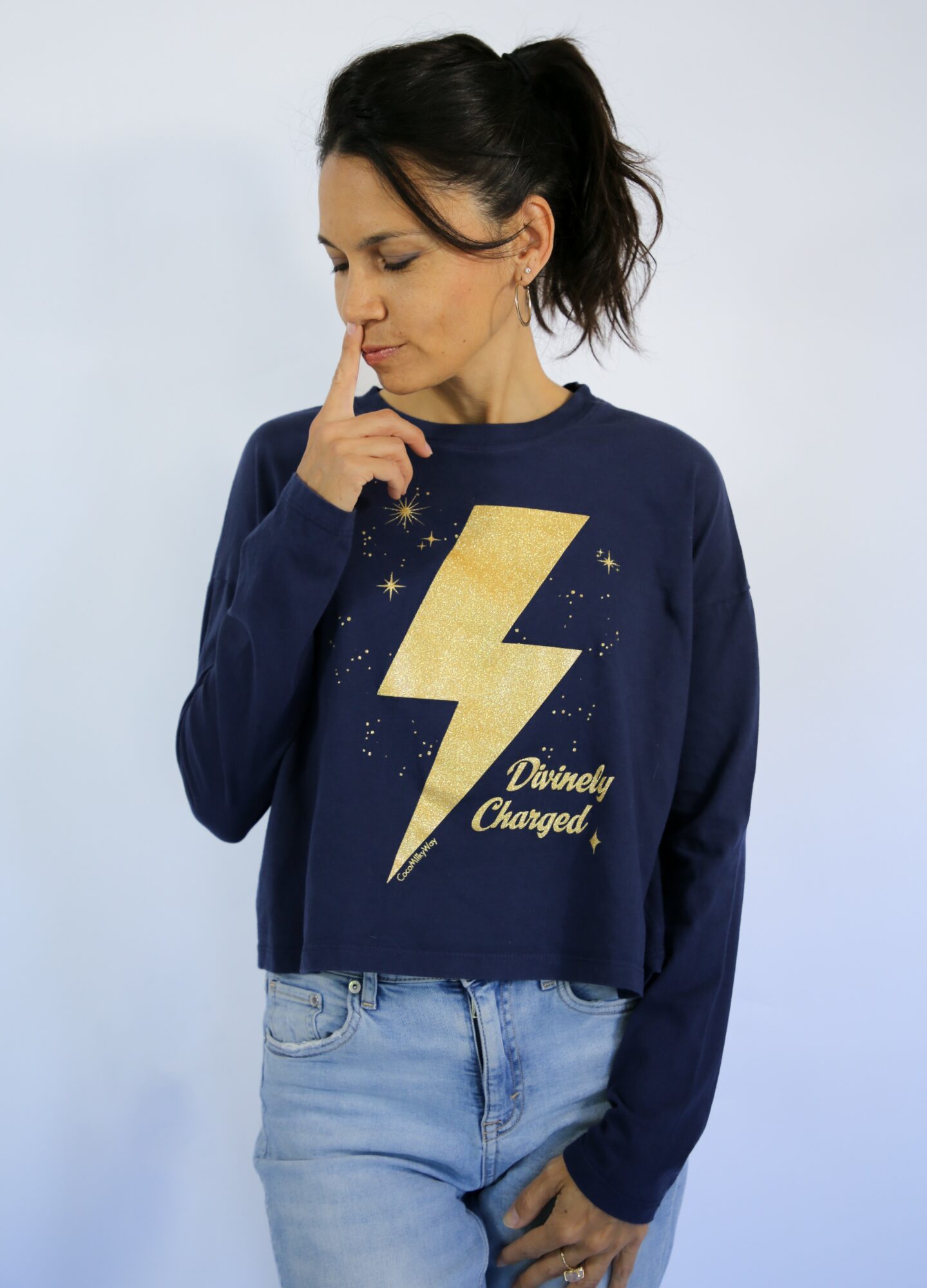 Woman with dark hair in a ponytail wearing a navy sweatshirt with a lightning bolt and text, looking down with finger on lips.