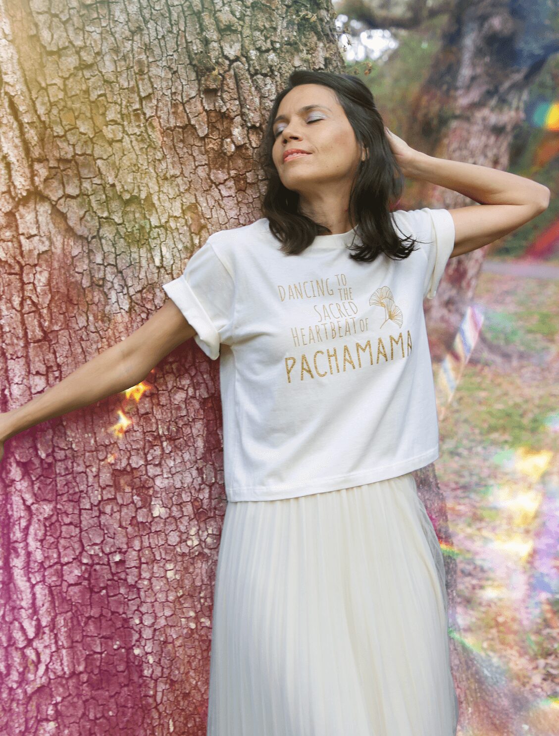 Woman leaning against a tree with eyes closed, wearing a white t-shirt and pleated skirt, outdoors in a park.