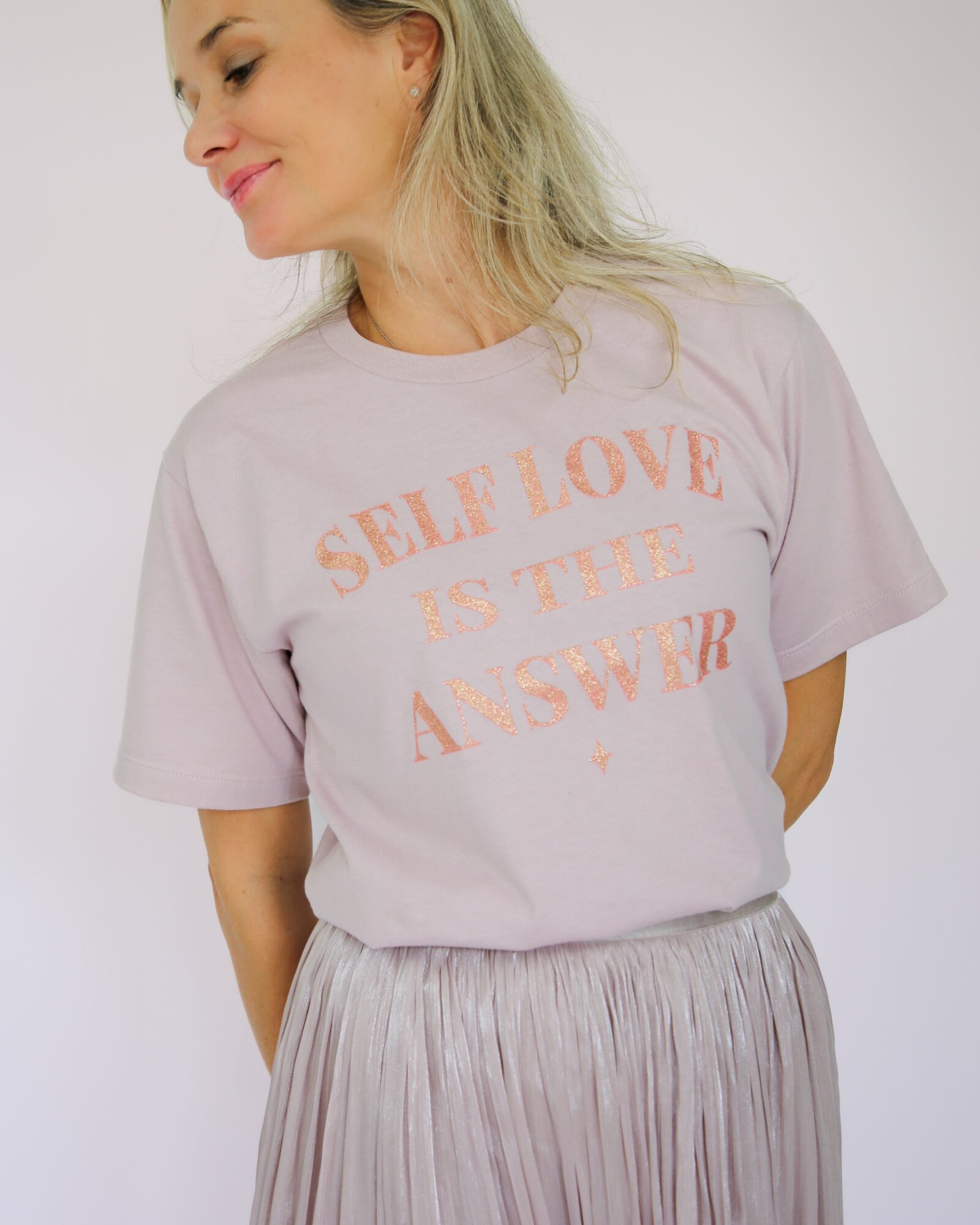 Woman with blonde hair wearing a light pink T-shirt with orange text and a pleated skirt, smiling sideways.