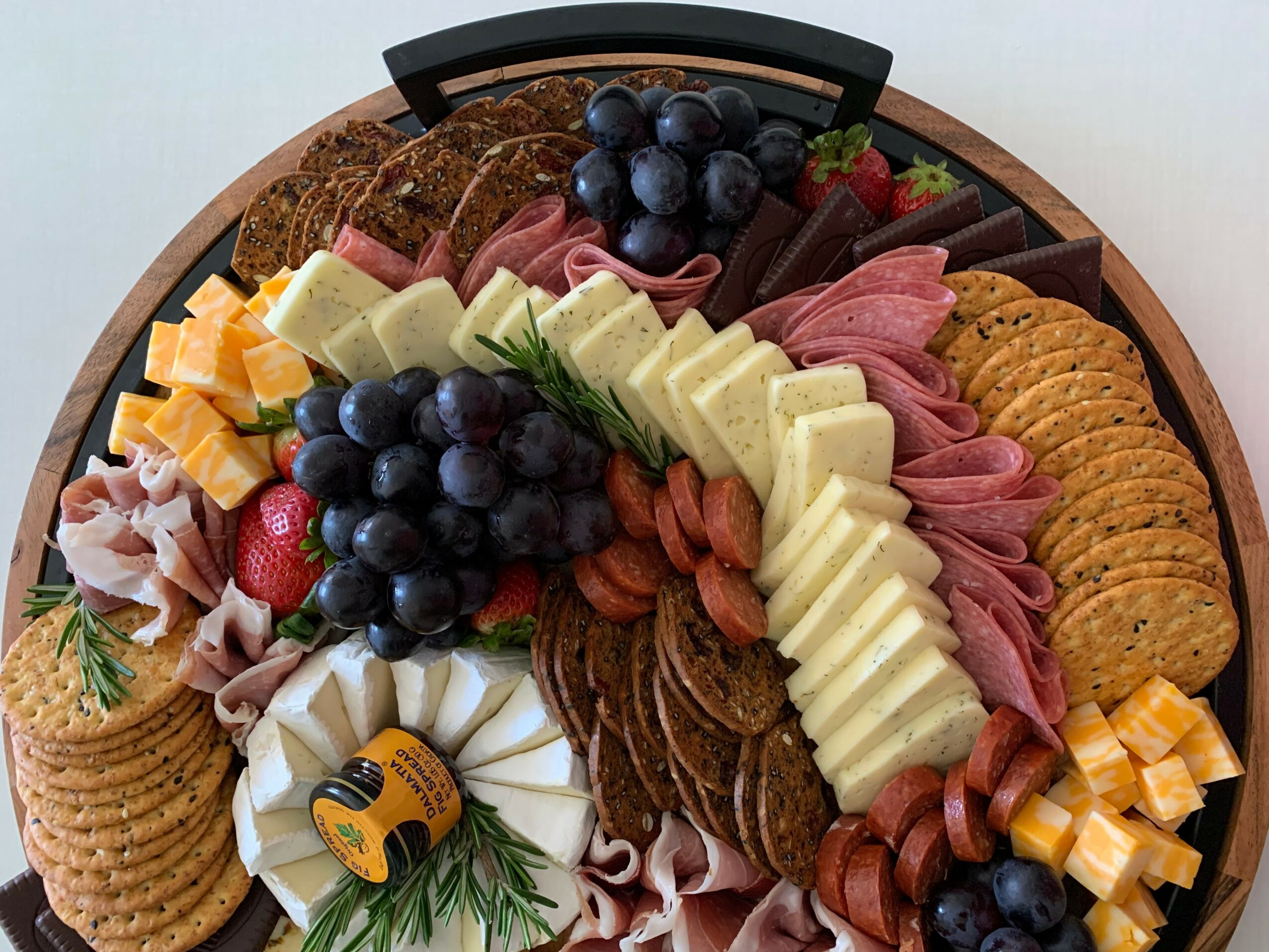 Round wooden platter with assorted cheeses, grapes, sliced meats, crackers, and strawberries, arranged in sections.
