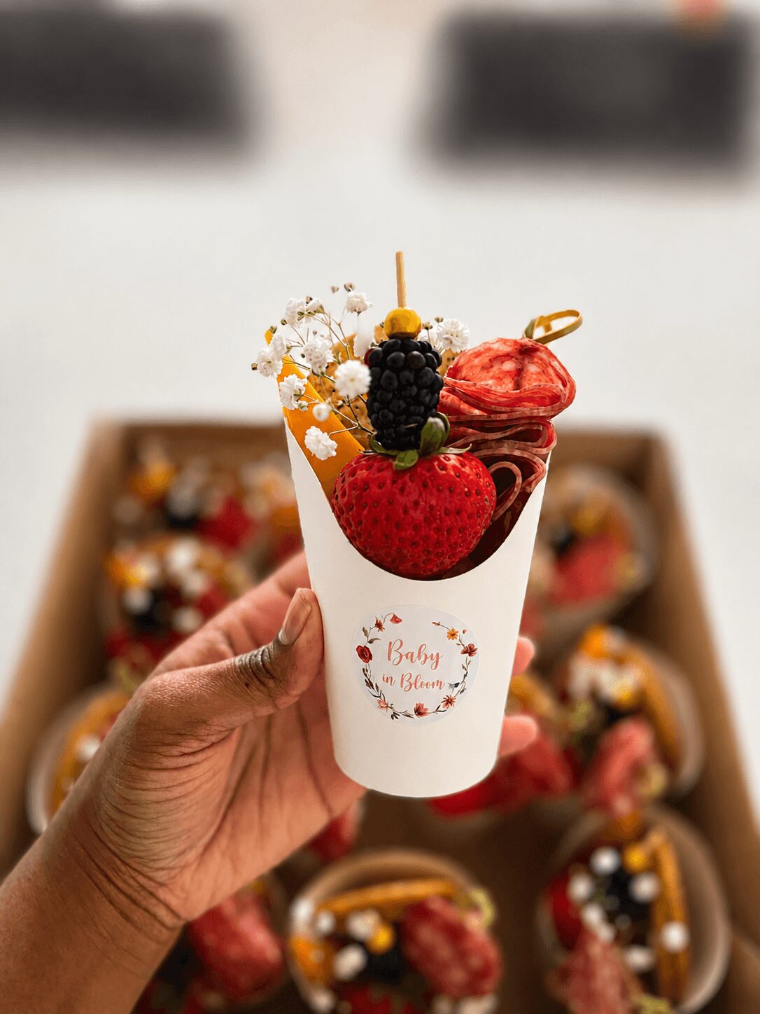 Hand holding a cup with assorted desserts, including a strawberry, blackberries, and decorative toppings, in front of a box of similar treats.