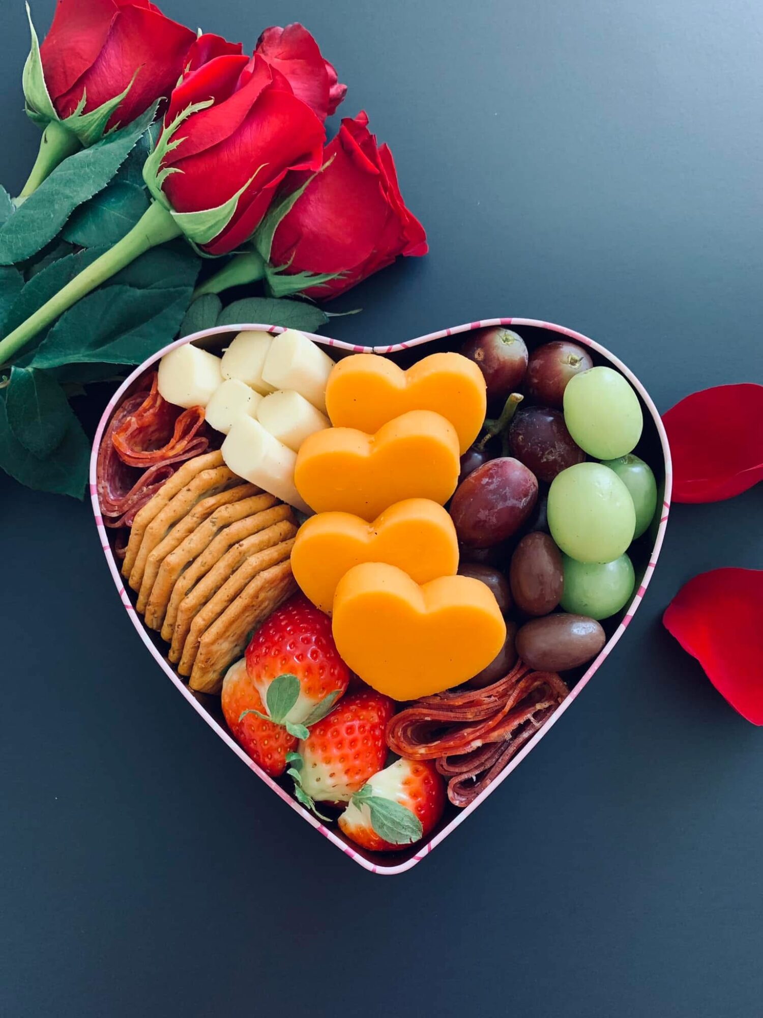Heart-shaped box filled with chocolates, strawberries, and cookies, with red roses and petals nearby.