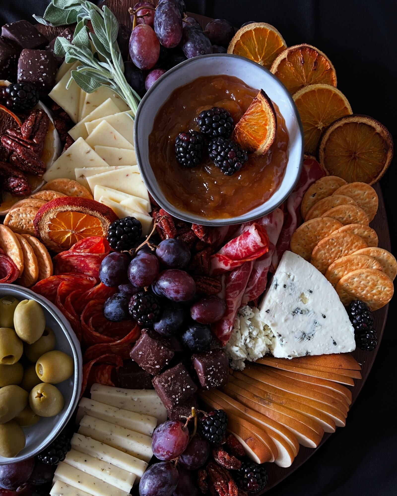 Assorted cheeses, fruits, and a bowl of jam on a platter, with slices of orange and other fruits, arranged for serving.