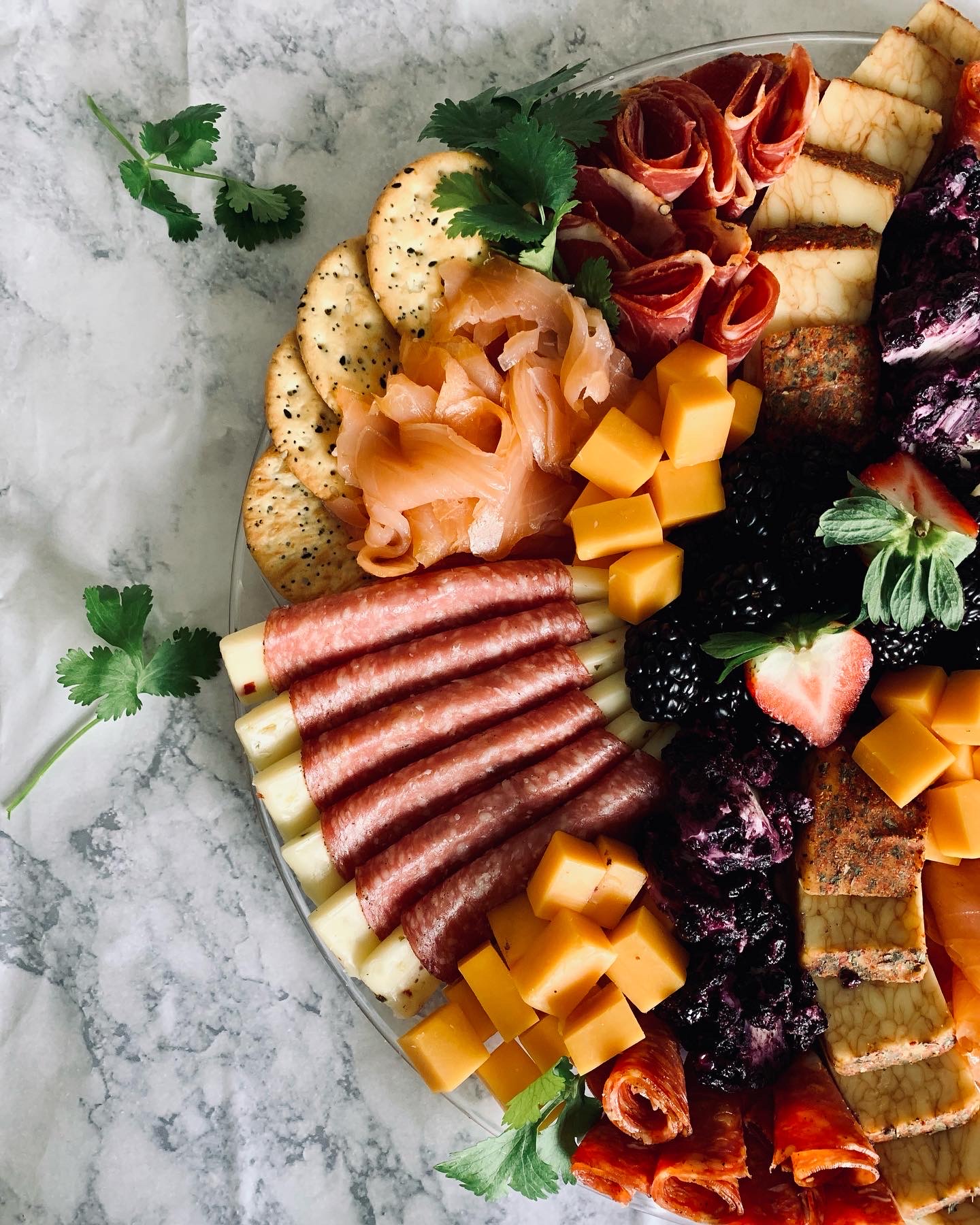 Assorted charcuterie board with sliced meats, cheeses, crackers, berries, and garnishes on a marble surface.