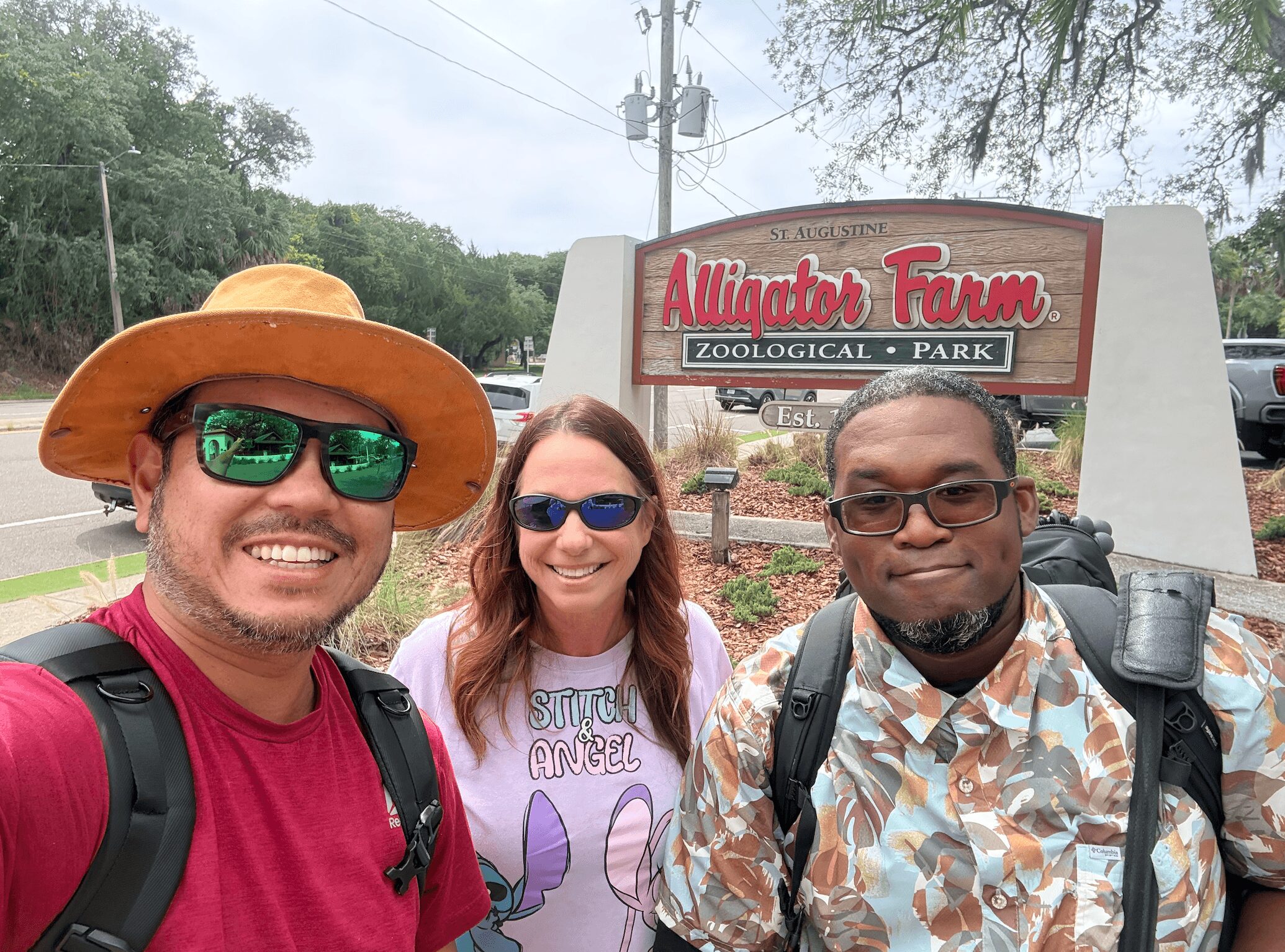 Three people smiling in front of a sign for Alligator Farm Zoological Park, with trees and a road in the background.