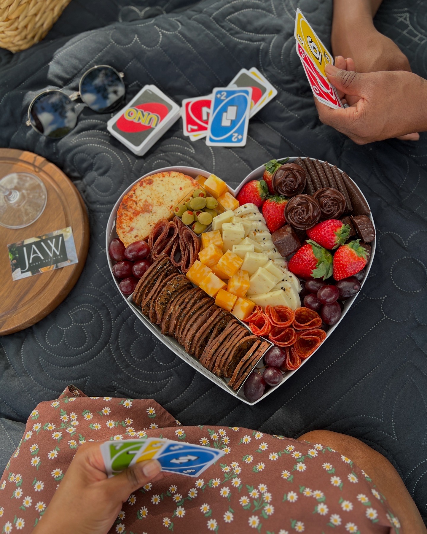 People playing card games with a heart-shaped box of assorted chocolates and strawberries on a dark surface.