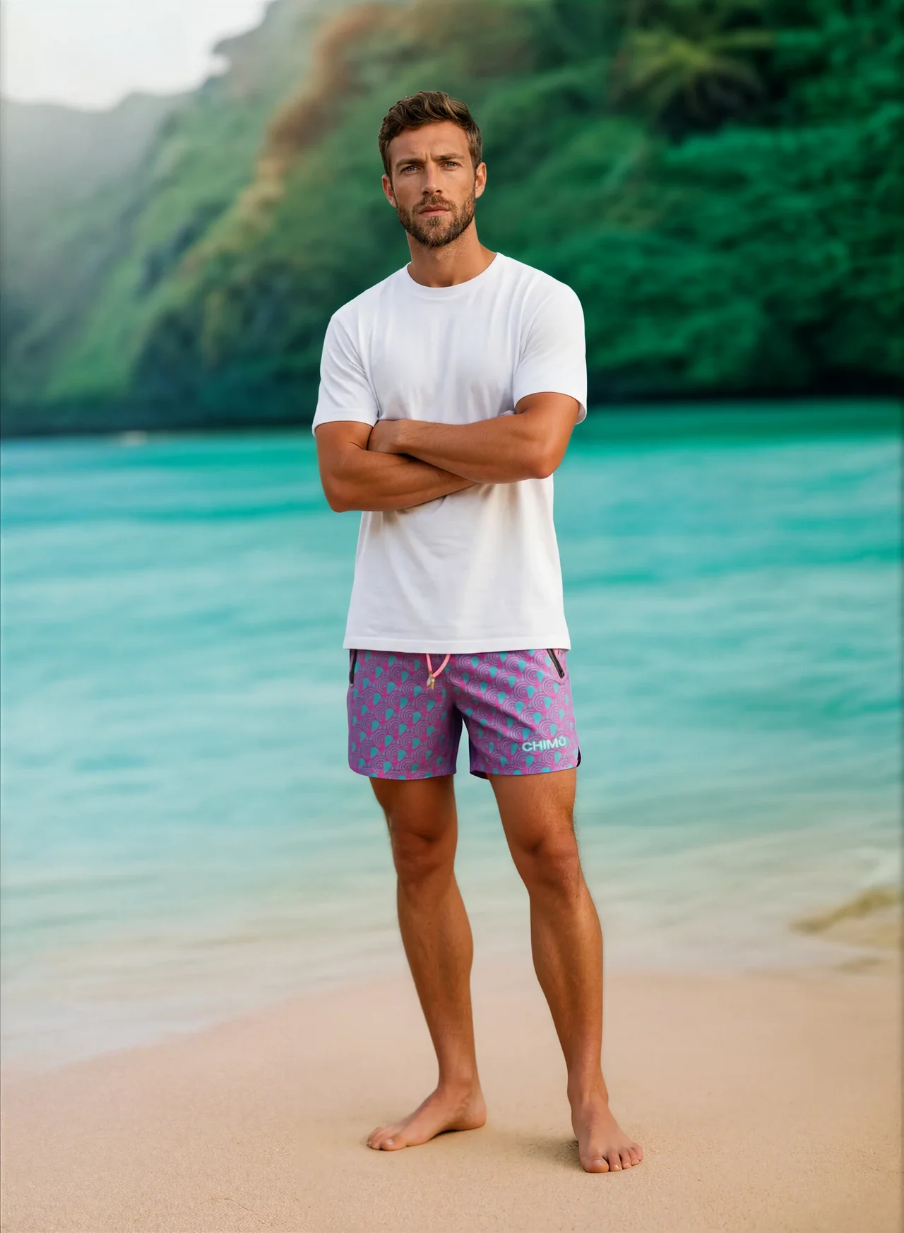 Man standing on beach with arms crossed, wearing white t-shirt and purple shorts, ocean and green hills in background.