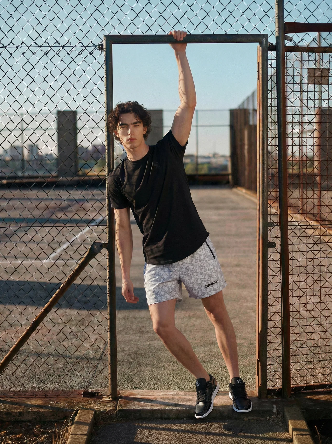 Young person standing in a gate opening, holding the top bar with one hand, outdoors on a sunny day.