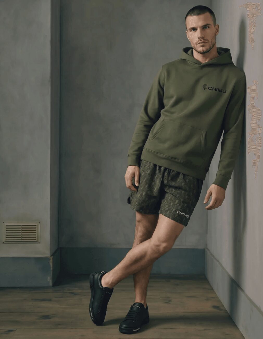 Man leaning against a wall wearing a green hoodie, black shorts, and black shoes in a room with gray walls.