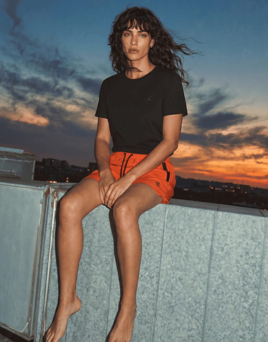 Woman sitting on a ledge during sunset, wearing a black t-shirt and orange shorts, with a cloudy sky background.