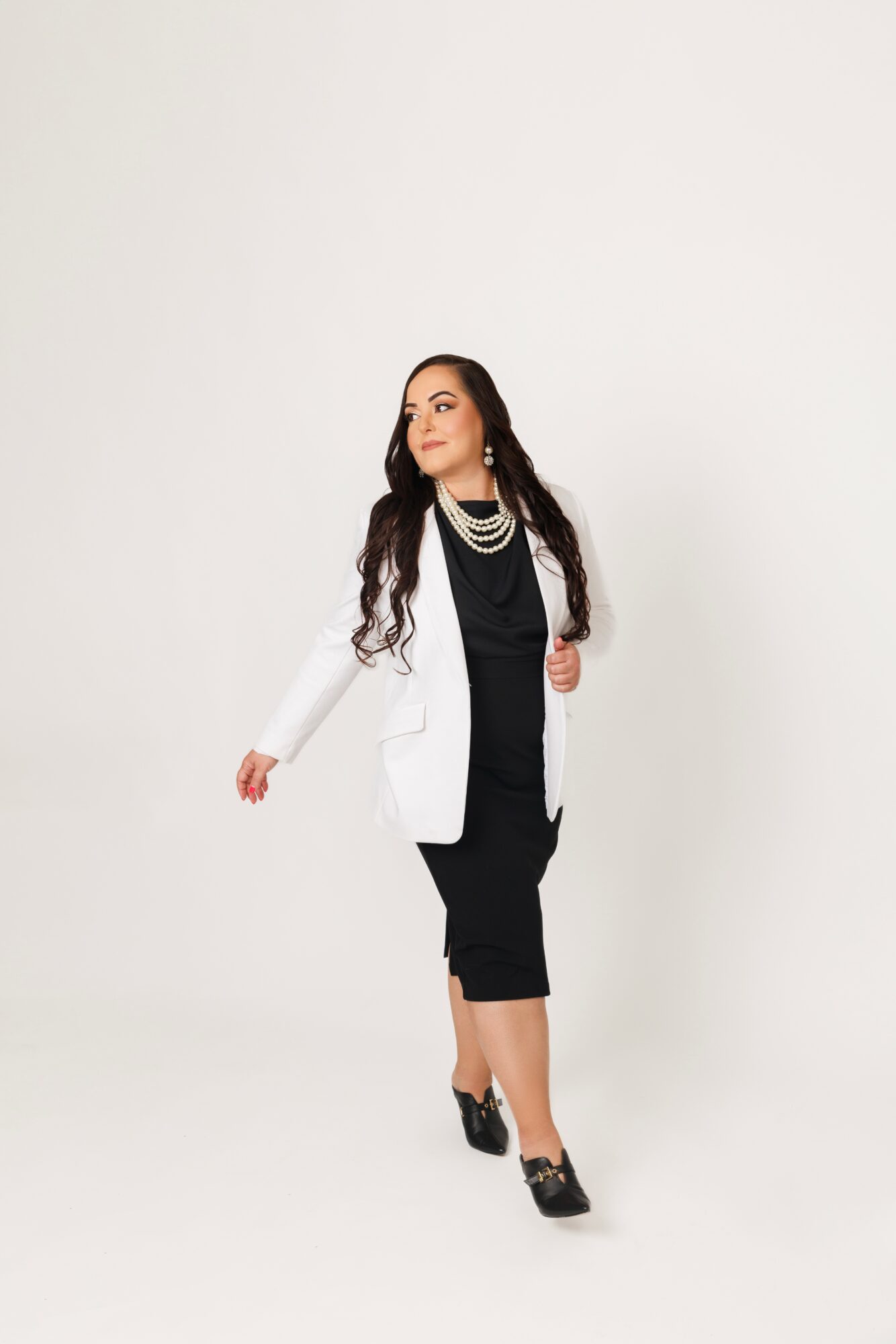 Woman in black dress and white blazer with jewelry, walking against a plain background.